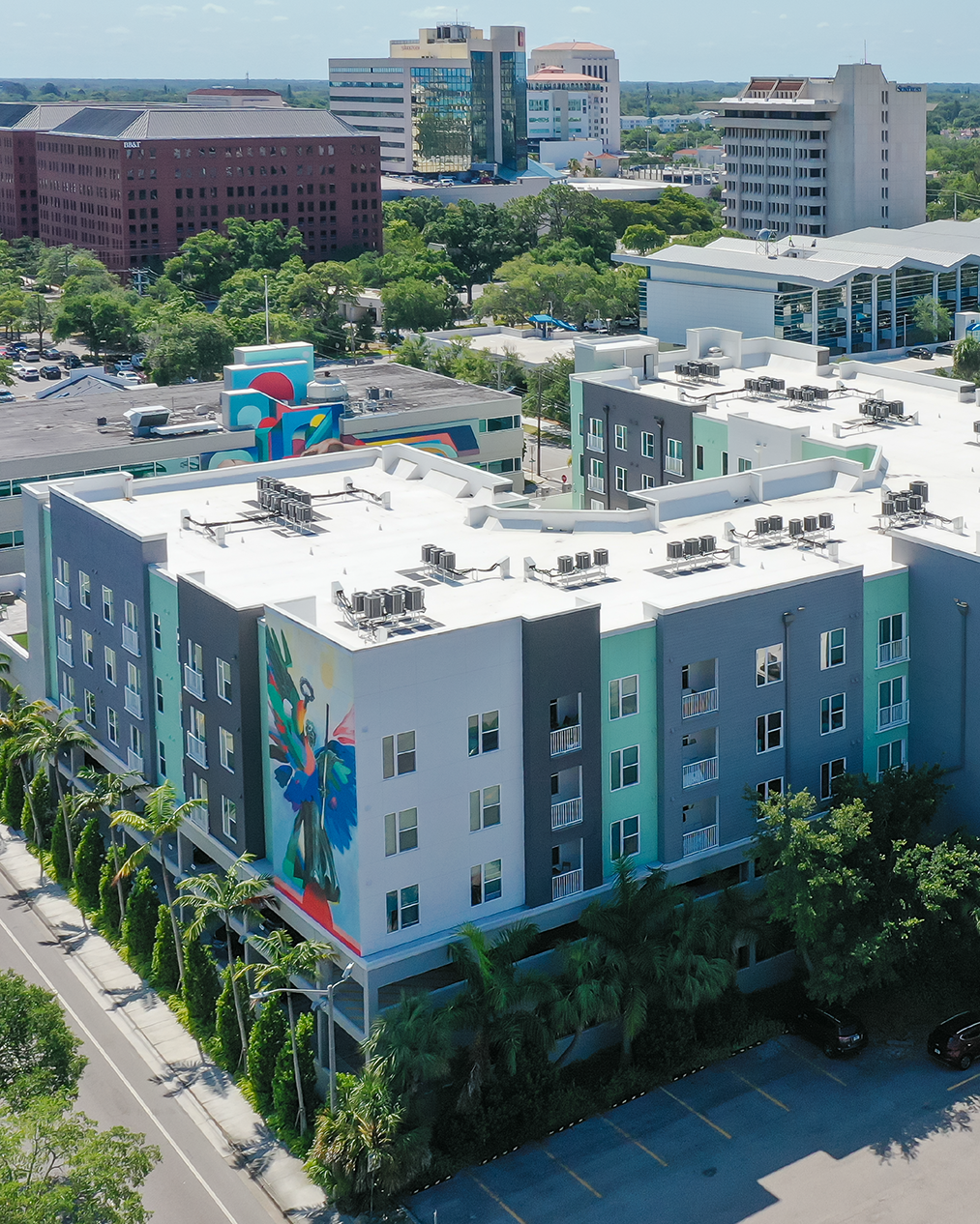 An aerial view of a modern apartment building featuring vibrant murals on the facade. Surrounding the building are palm trees and a busy urban landscape with various structures in the background.