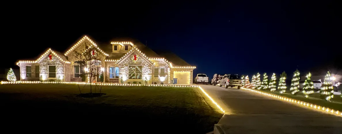 Wide view of a large home lit in warm white across every roofline and window, with wreaths on the gables and a lit path of shrubs running the length of the driveway.