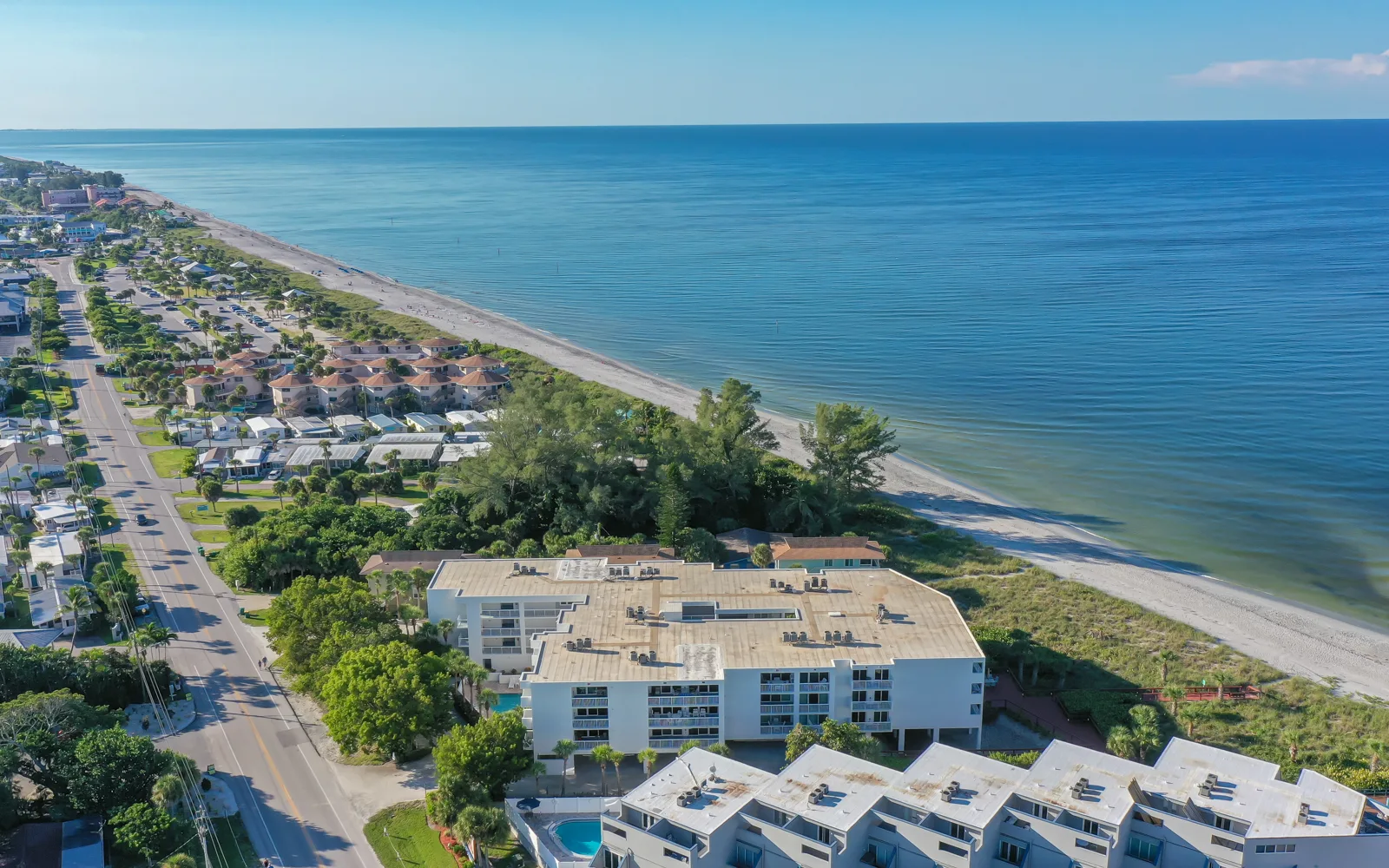 An aerial view of a coastal area featuring sandy beaches alongside residential buildings and lush greenery. The calm blue ocean extends into the horizon under a clear sky.