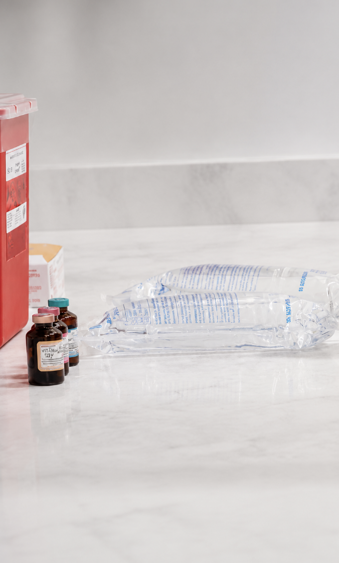 Medical supplies including saline solution bags, a biohazard disposal container, and glass vials of medication on a marble countertop.