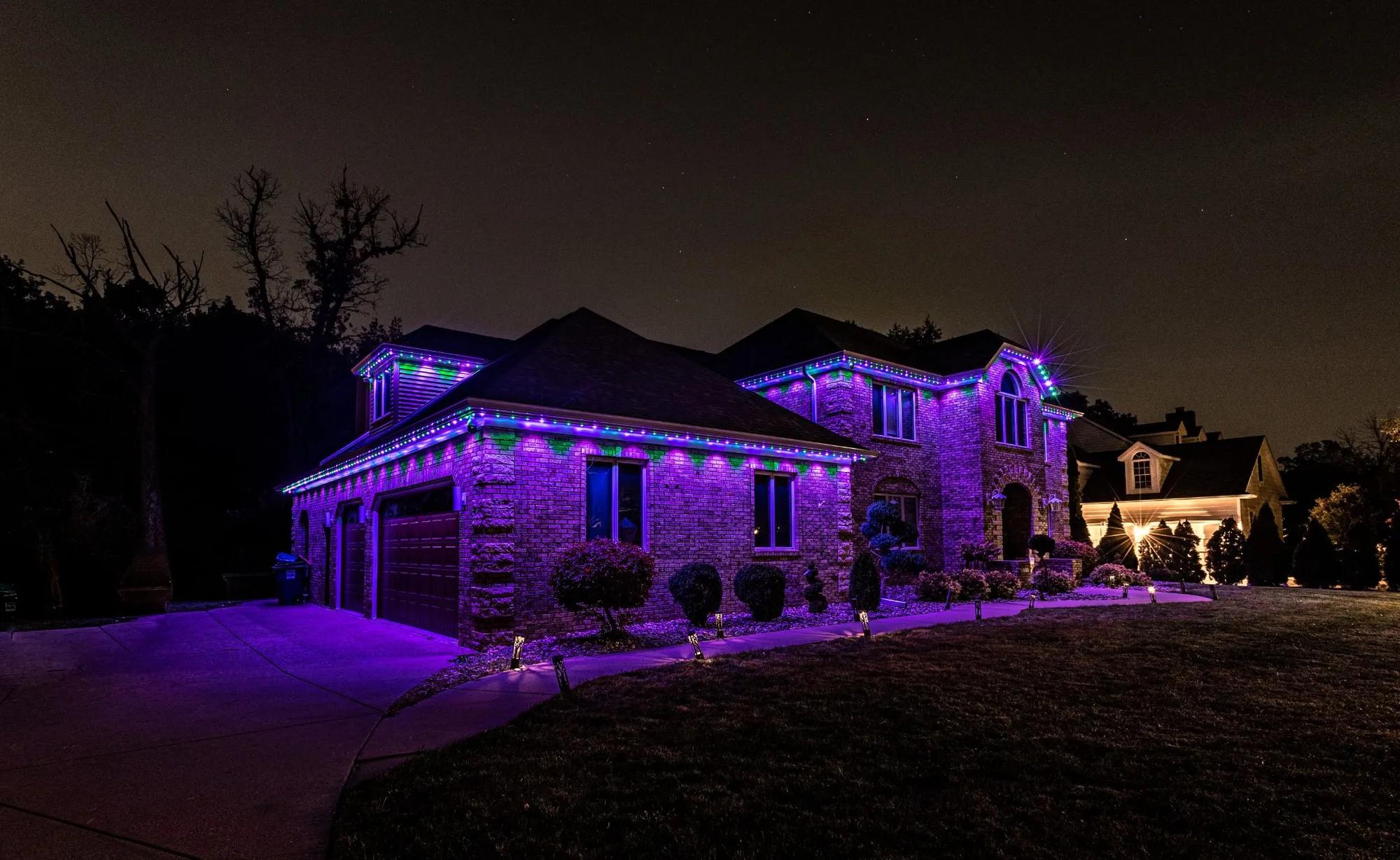 The same brick home with its permanent lighting switched to a purple and green scene, washing the brickwork in colour while the roofline picks out the eaves.