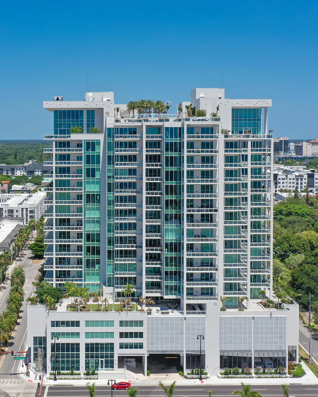 This image features a contemporary high-rise building with a glass and concrete facade. It includes several floors of residential units and landscaped terraces, set against a clear blue sky.