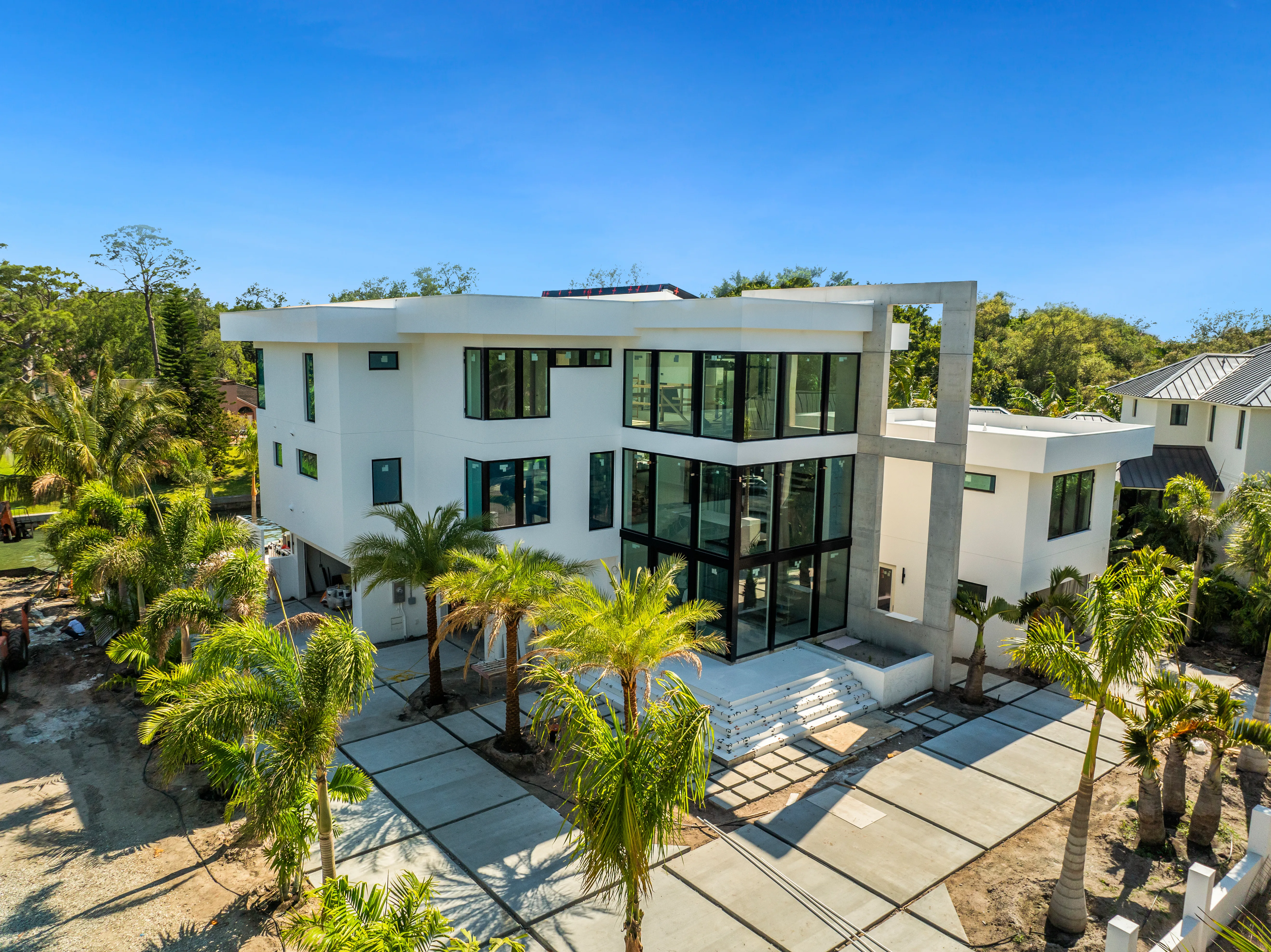 A contemporary multi-story house featuring large windows and a sleek design, surrounded by palm trees. The property includes a spacious driveway and is set against a clear blue sky.