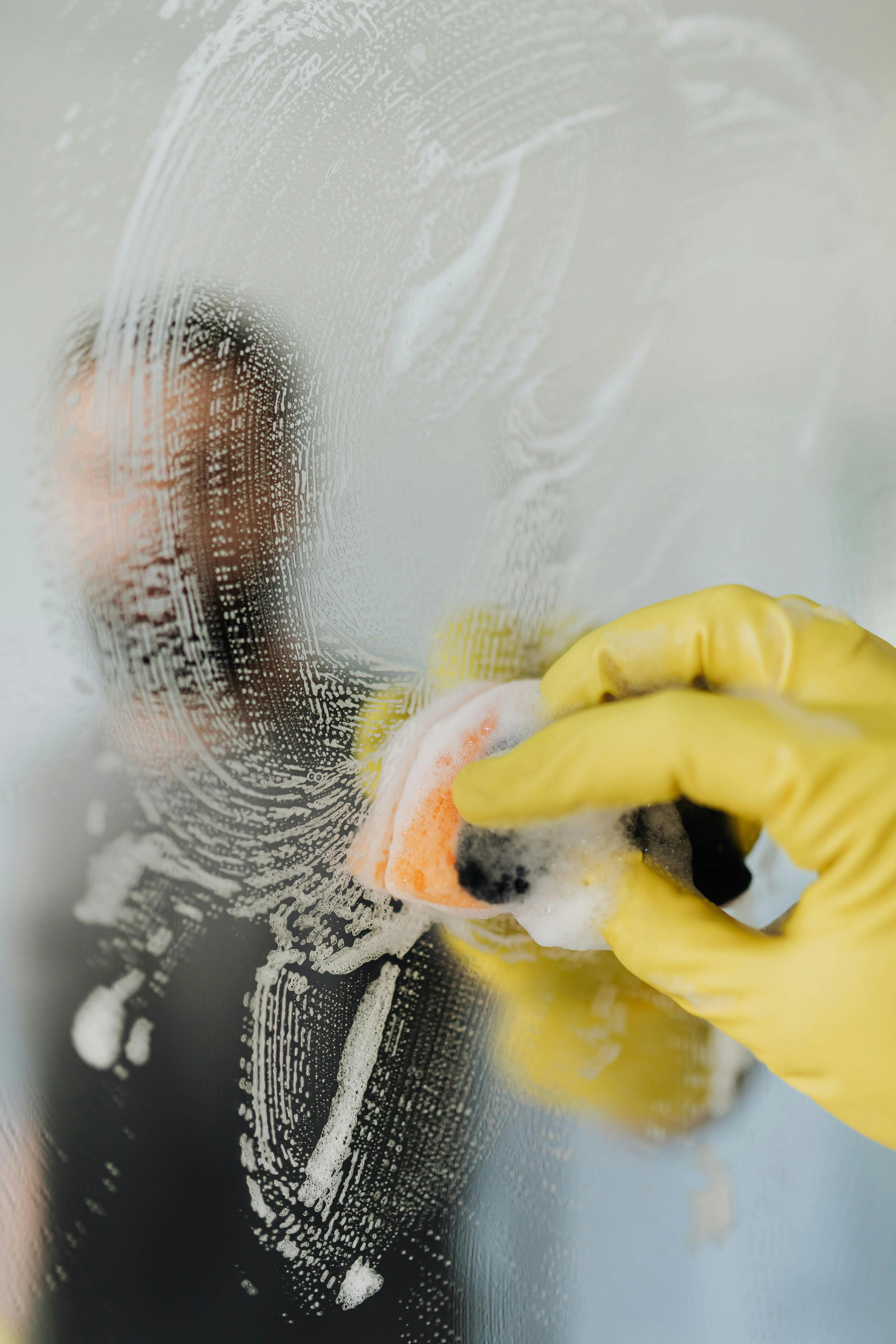 Person wearing yellow gloves cleaning a glass surface with a soapy sponge