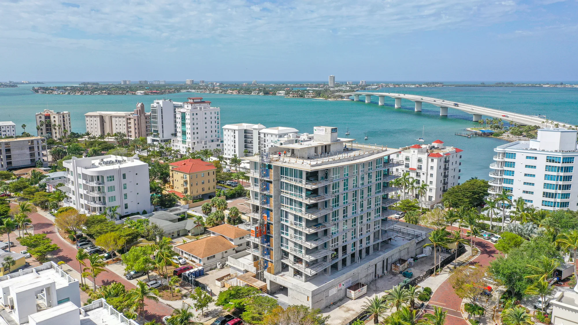 An aerial view of a coastal city featuring a mix of residential buildings and an under-construction high-rise. The turquoise waters and a bridge connecting landmasses are visible, with palm trees lining the streets.