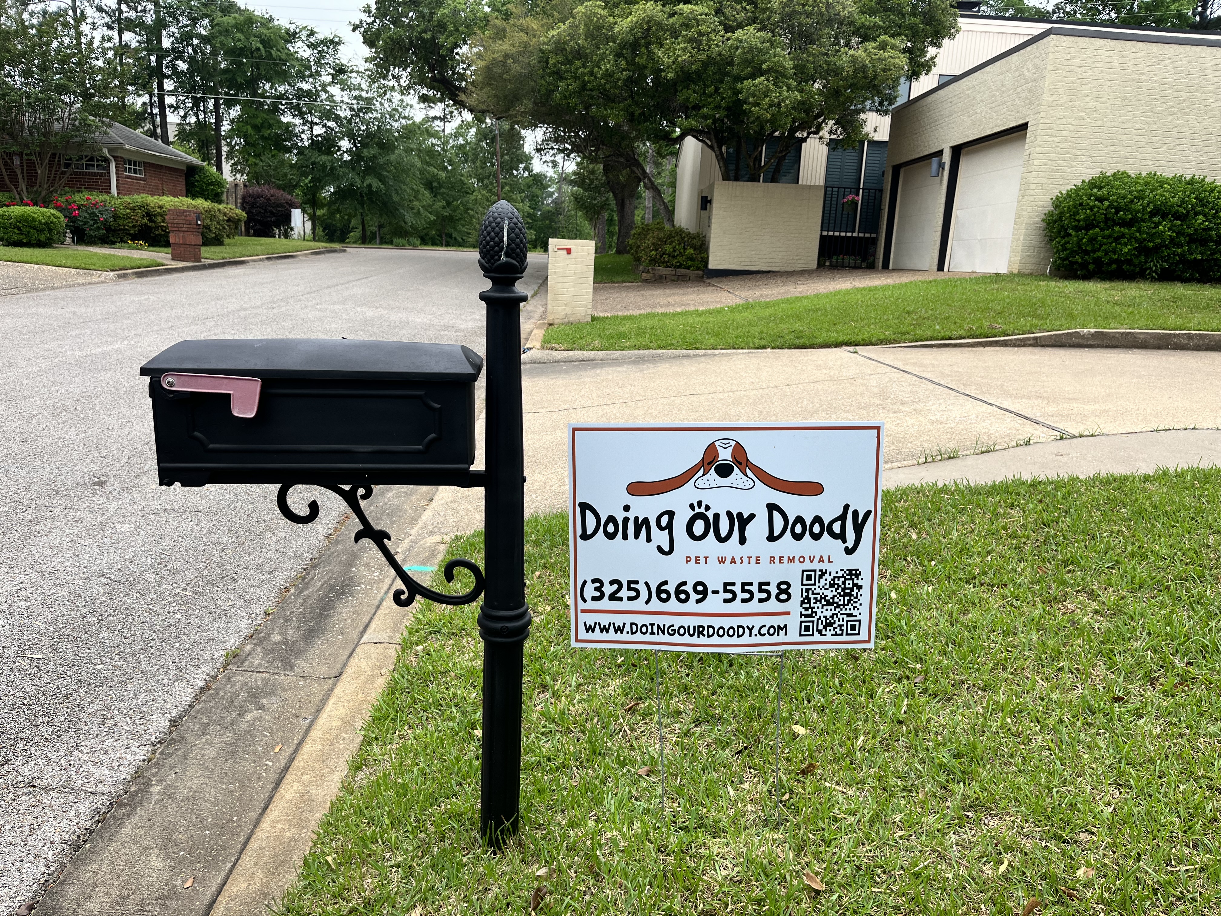 Doing Our Doody yard sign with QR code beside a black mailbox on a suburban street