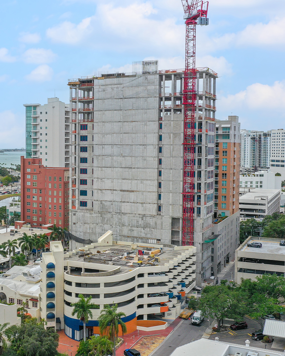 A partially completed high-rise building under construction, featuring a large crane, stands next to an existing multi-level parking structure. Various modern buildings are visible in the background, set against a cloudy sky.