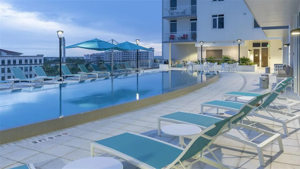 This image showcases a modern rooftop infinity pool surrounded by lounge chairs and umbrellas, with a view of buildings in the distance. The setting is illuminated by outdoor lamps, creating a serene atmosphere during twilight.