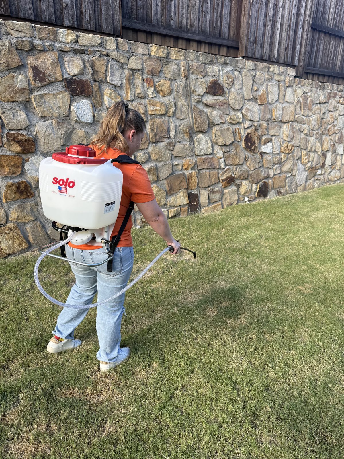 A person wearing an orange shirt and jeans operating a backpack sprayer in a yard with a stone wall backdrop