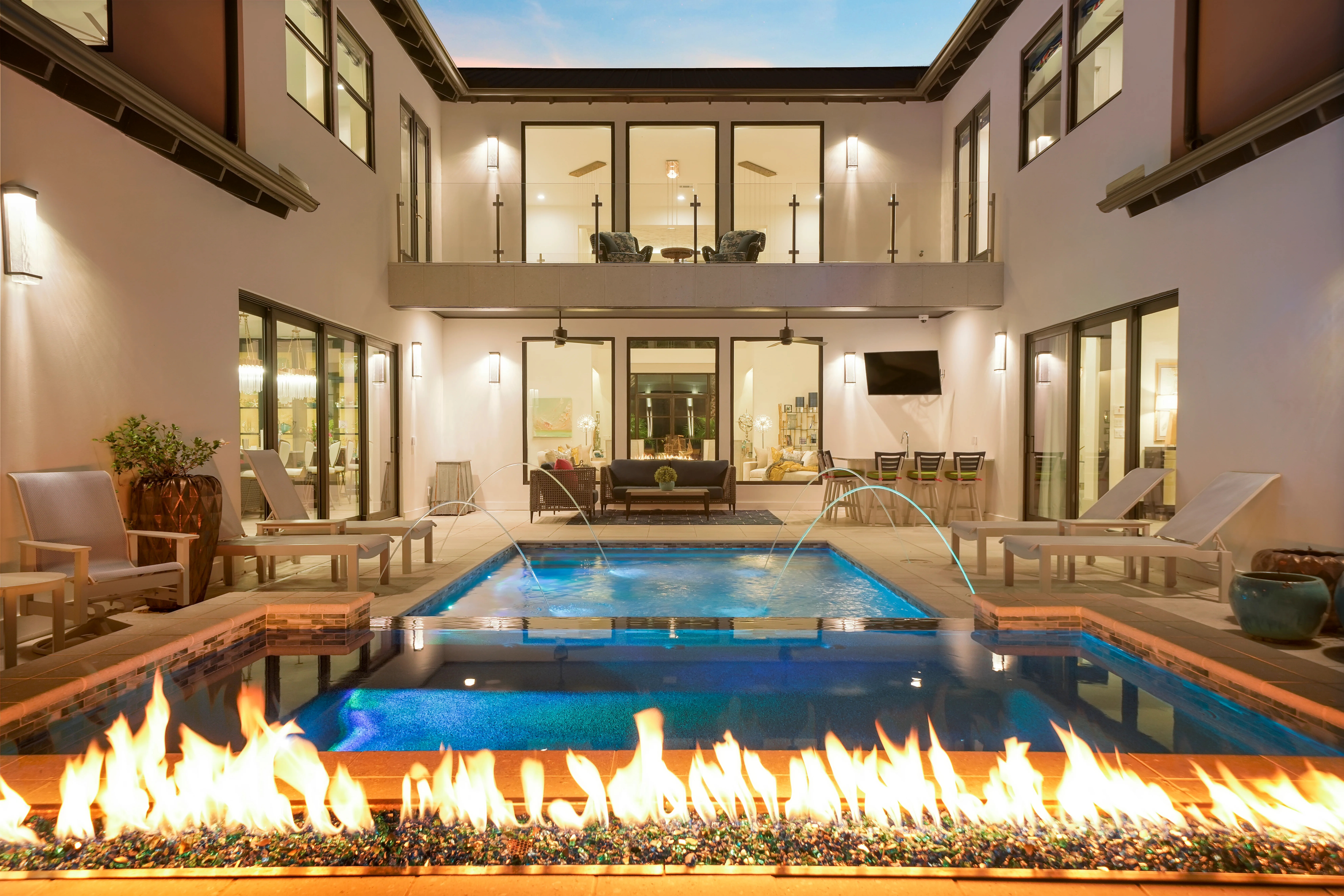 A swimming pool beside a long lit fire feature on a paved terrace at dusk.