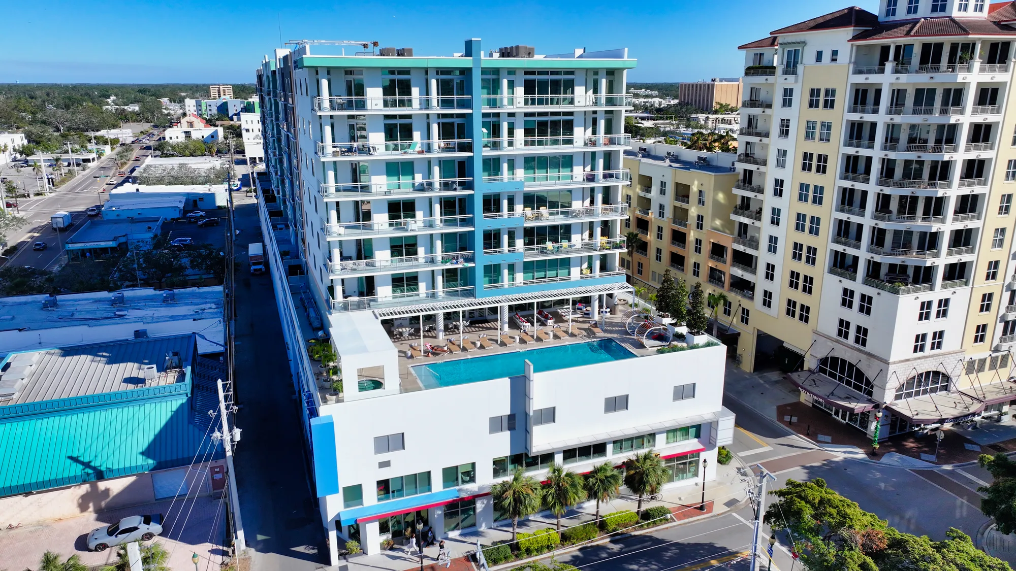 A modern multi-story apartment building featuring a rooftop pool and spacious balconies. Surrounding structures include nearby businesses and residential buildings, set against a clear blue sky.
