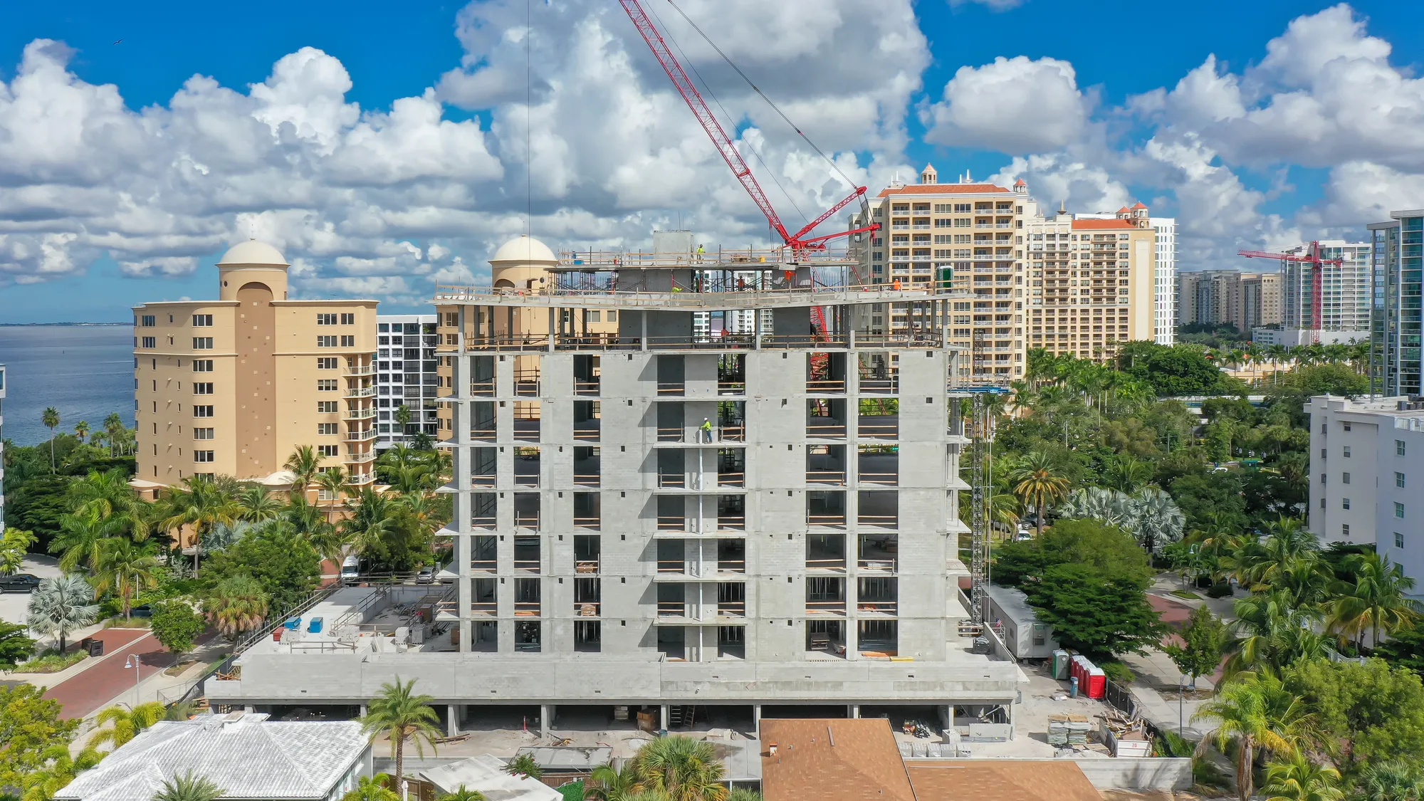 A partially constructed high-rise building dominates the foreground, with cranes and construction workers visible on the site. Surrounding the building are residential structures and lush greenery, with a body of water in the background under a bright blue sky filled with clouds.