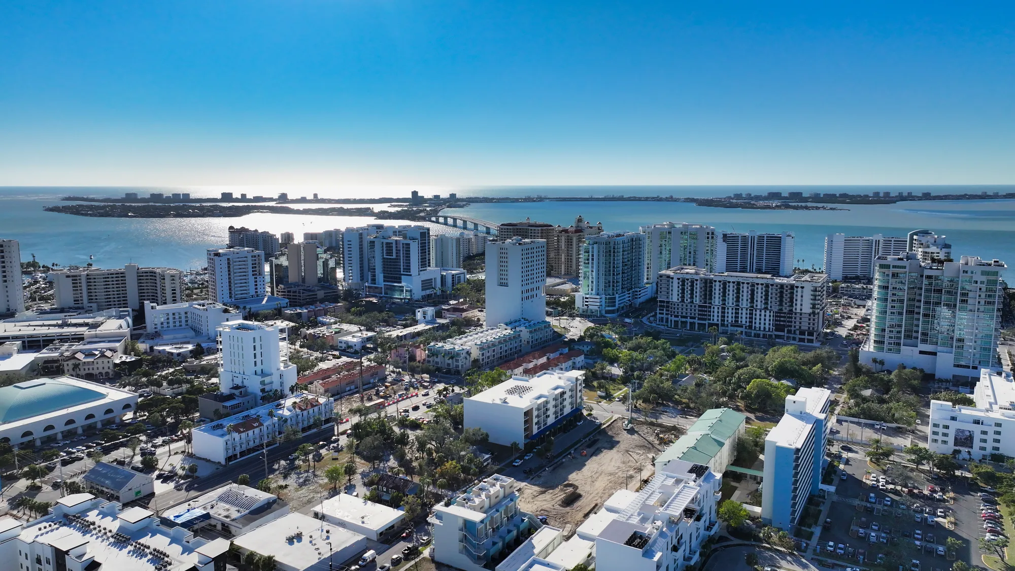 This aerial image showcases a modern waterfront city with high-rise buildings and a scenic body of water in the background. The bright blue sky and reflection on the water enhance the vibrant, urban atmosphere.