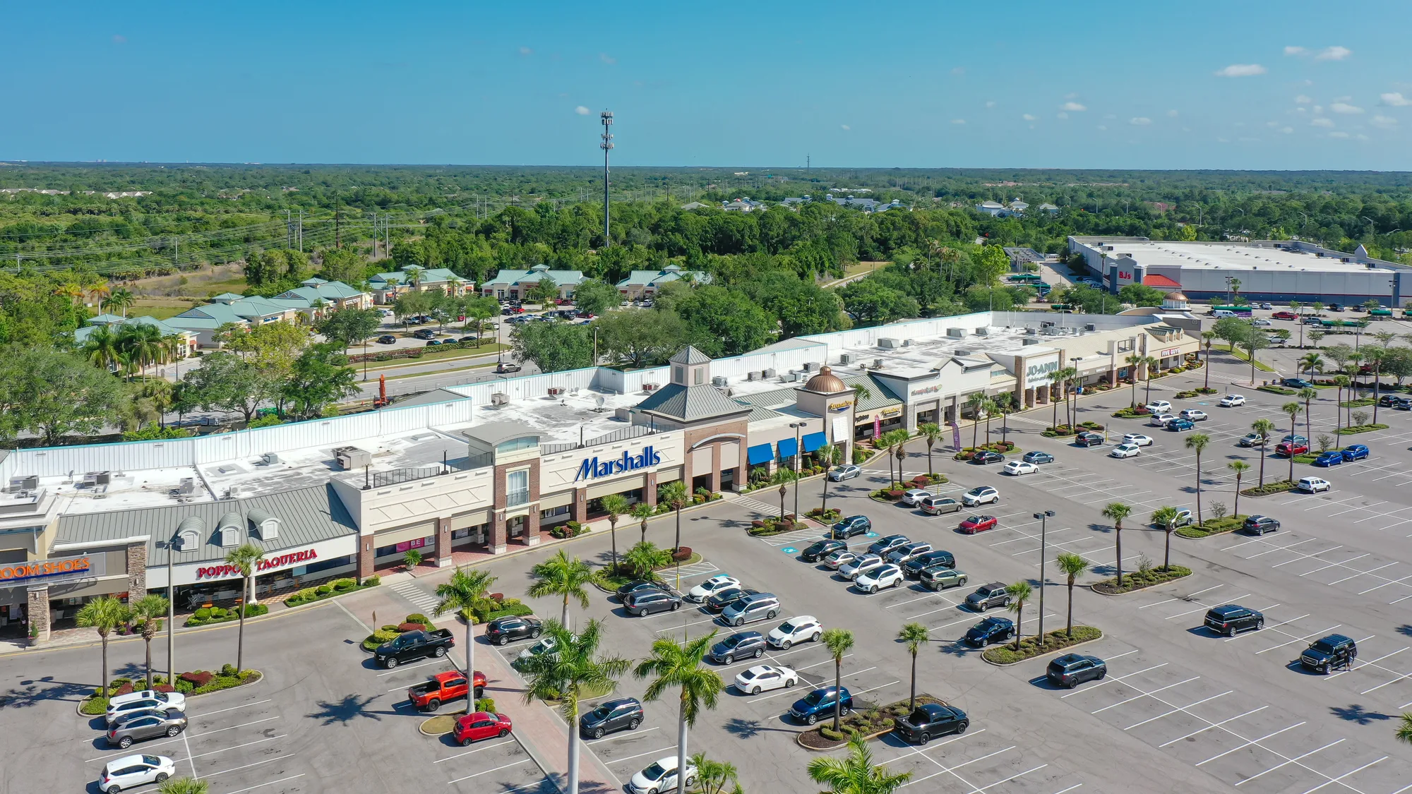 This aerial image features a shopping center with various retail stores, including Marshalls and Joann Fabrics, surrounded by a large parking lot. Palm trees line the parking area under a clear blue sky, with additional greenery and buildings visible in the background.