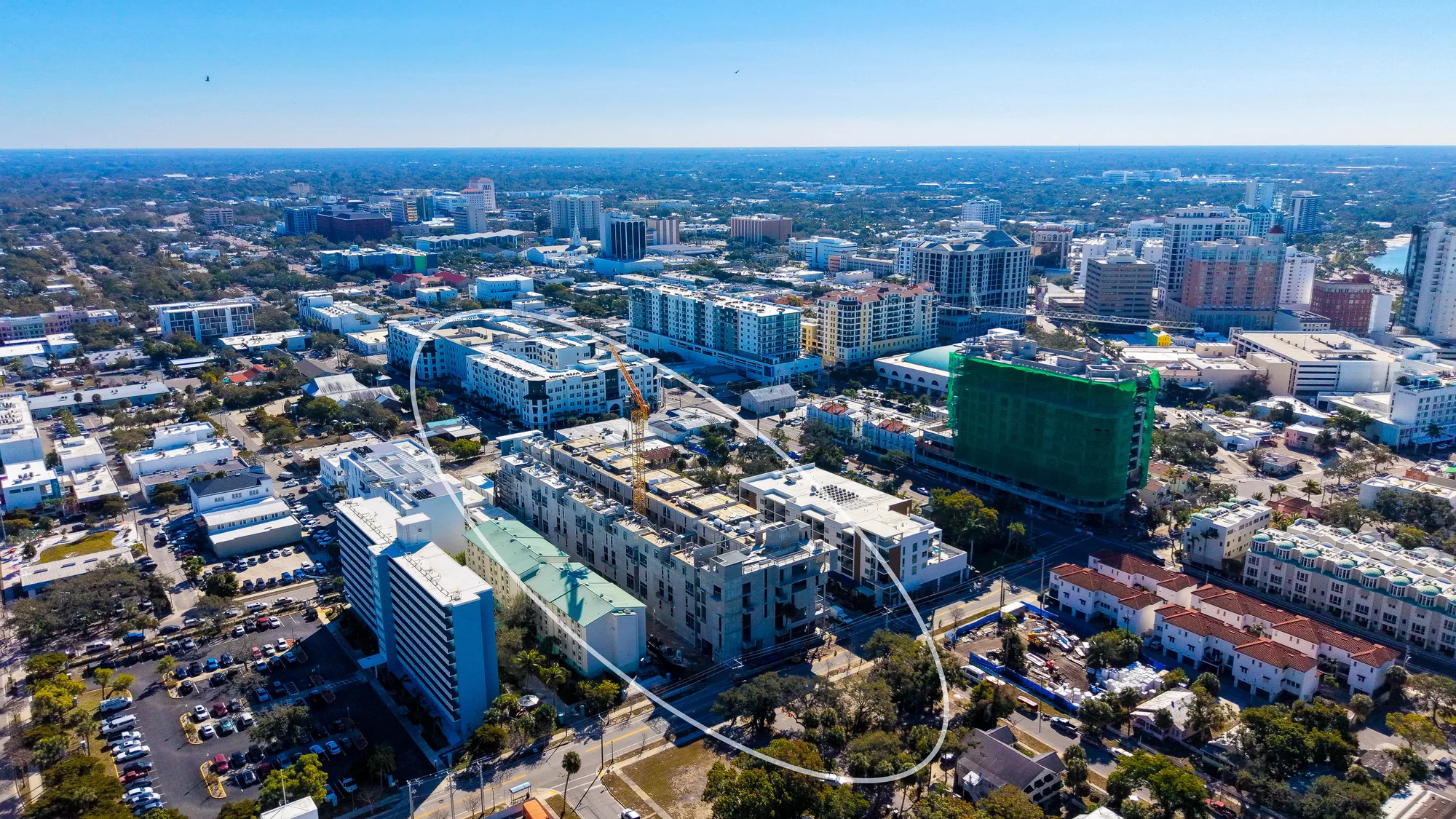 This aerial image captures a bustling urban landscape featuring a mix of commercial and residential buildings, with new construction evident in the foreground. The backdrop shows a city skyline under clear blue skies.