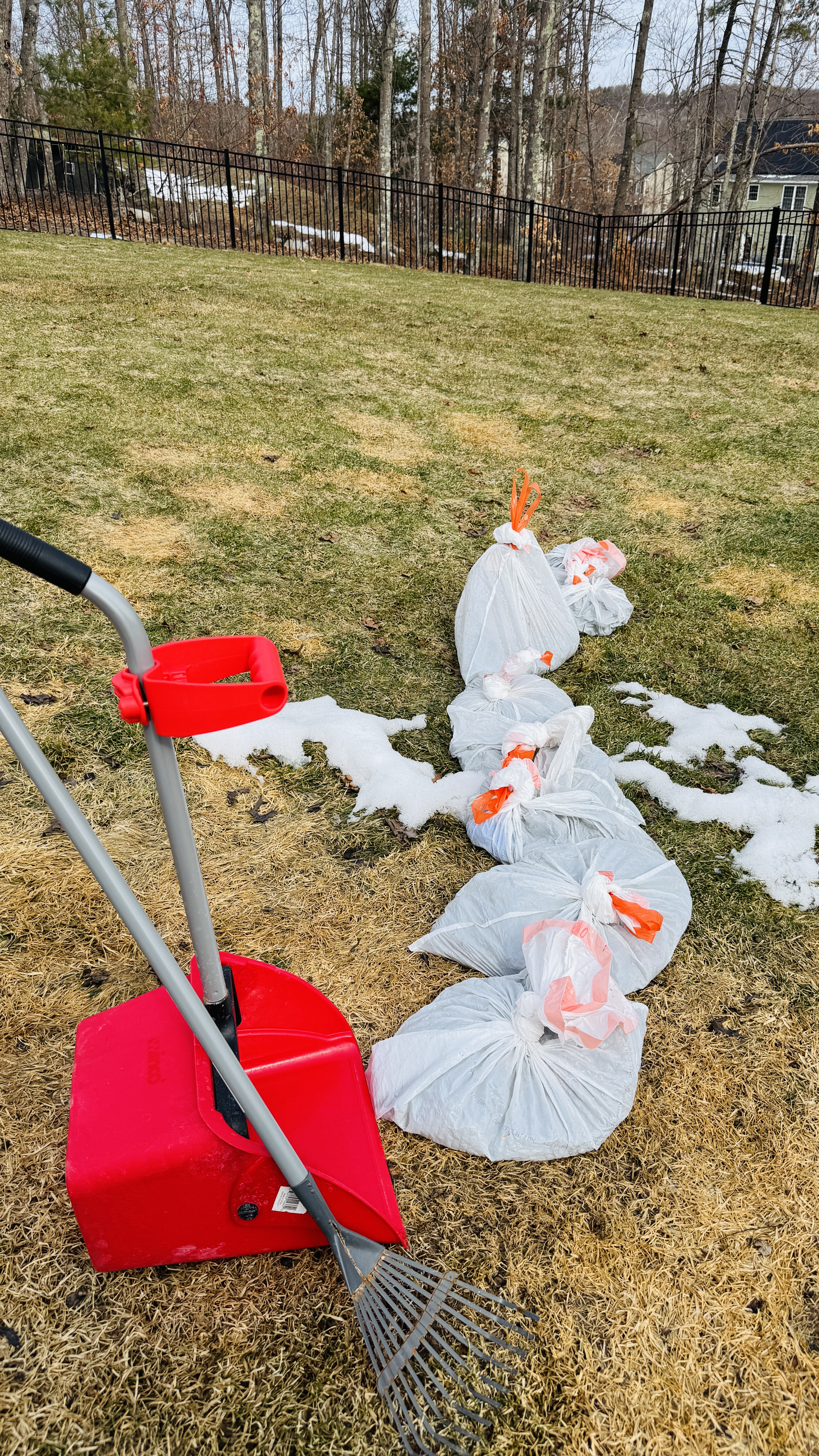 Red shovel and metal rake resting beside filled bags on thawing spring grass