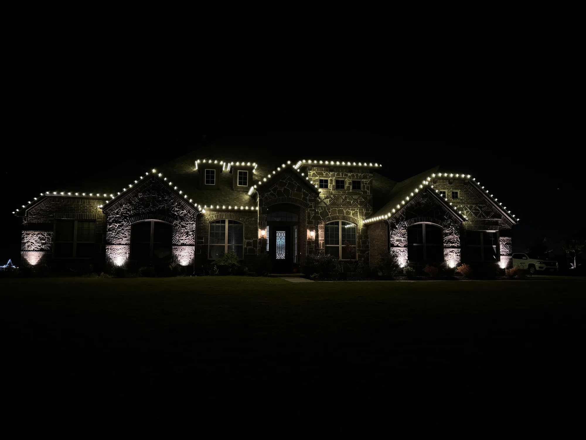 Large stone and brick home with warm white C9 bulbs following the full roofline, lit by landscape uplighting below.