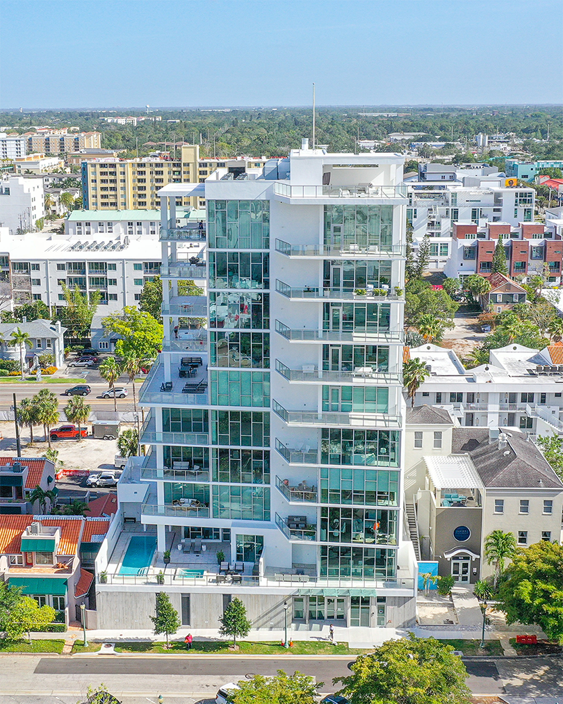 A contemporary glass high-rise building features multiple balconies and a swimming pool on the lower level. Surrounded by residential areas and cityscape, the building showcases modern architecture against a clear blue sky.