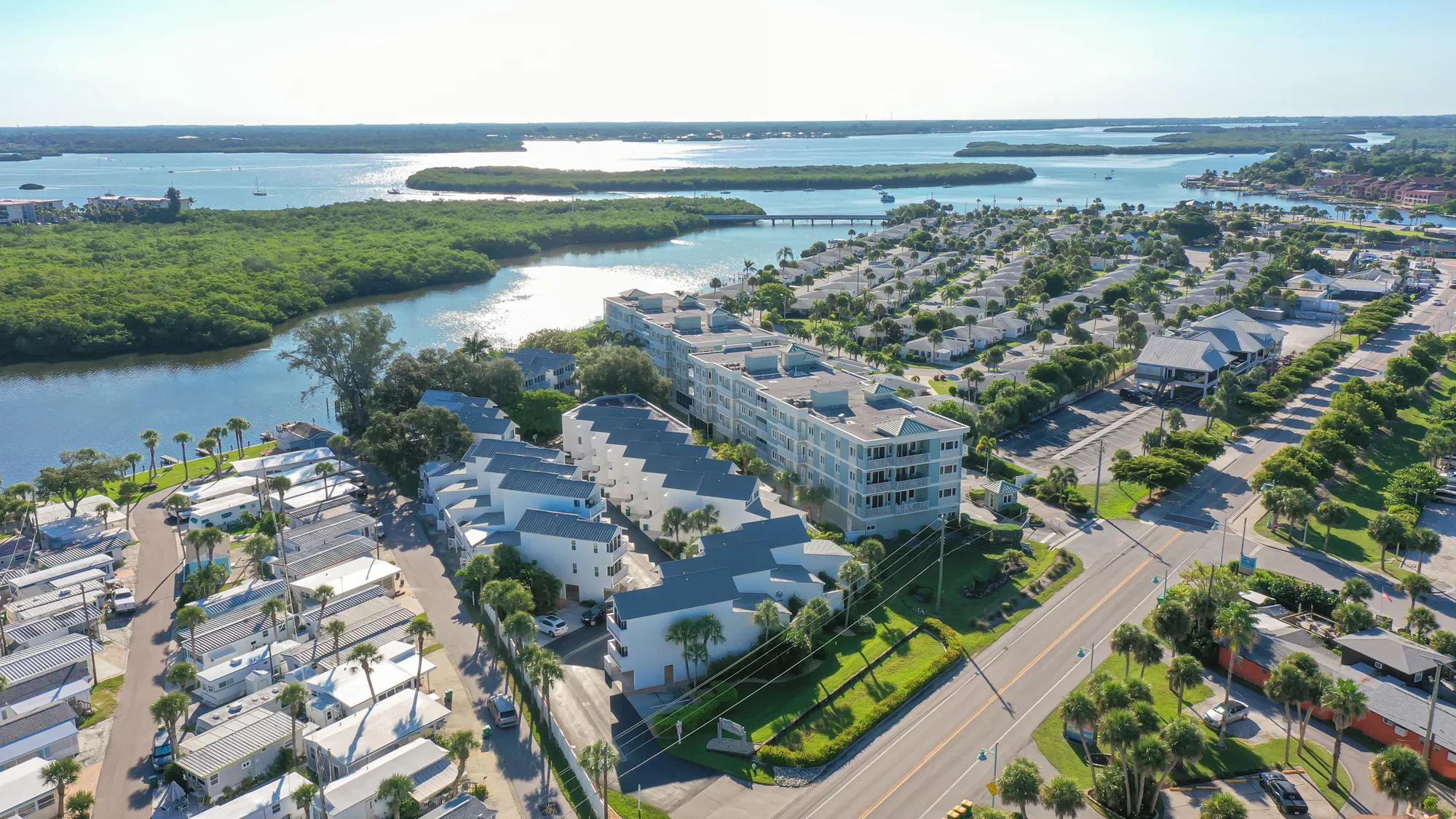 An aerial image captures a coastal community featuring multi-story buildings and smaller homes along a waterfront. The scene includes a river, surrounding greenery, and a neighborhood with structured roads and parked vehicles.