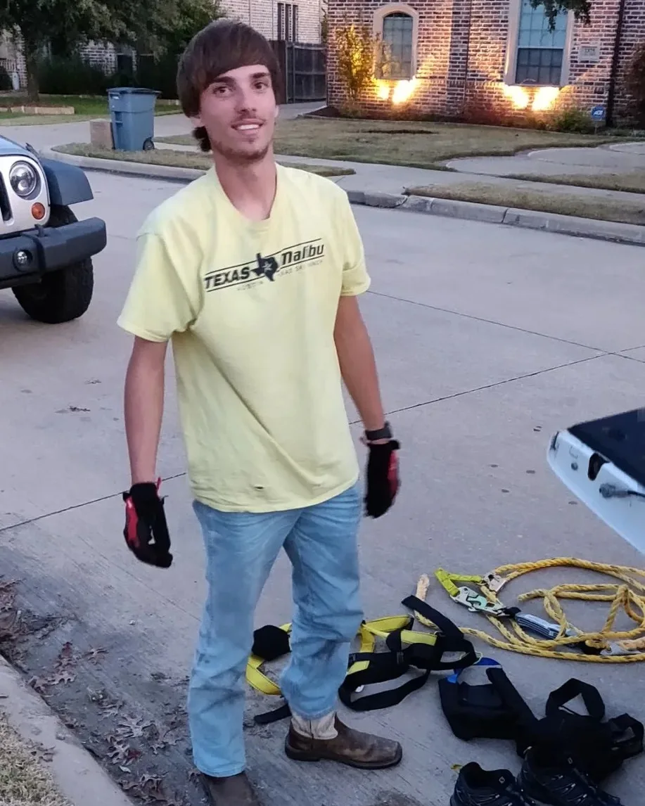 Installer standing on a residential street beside coiled rope and safety harnesses, with a lit home in the background.