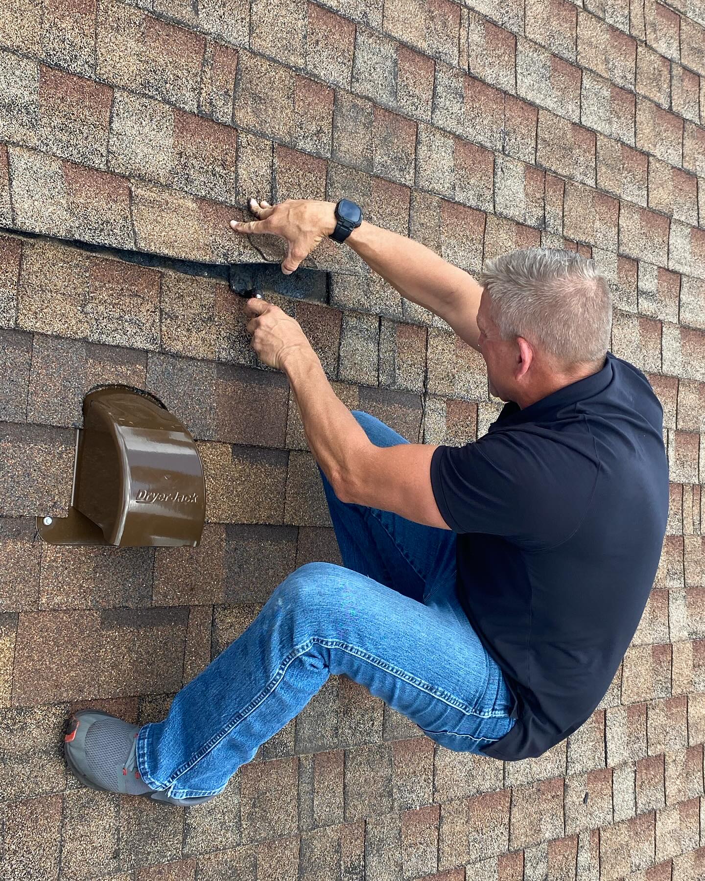 A man installing a dryer vent on a sloped roof, securing hardware on the shingles