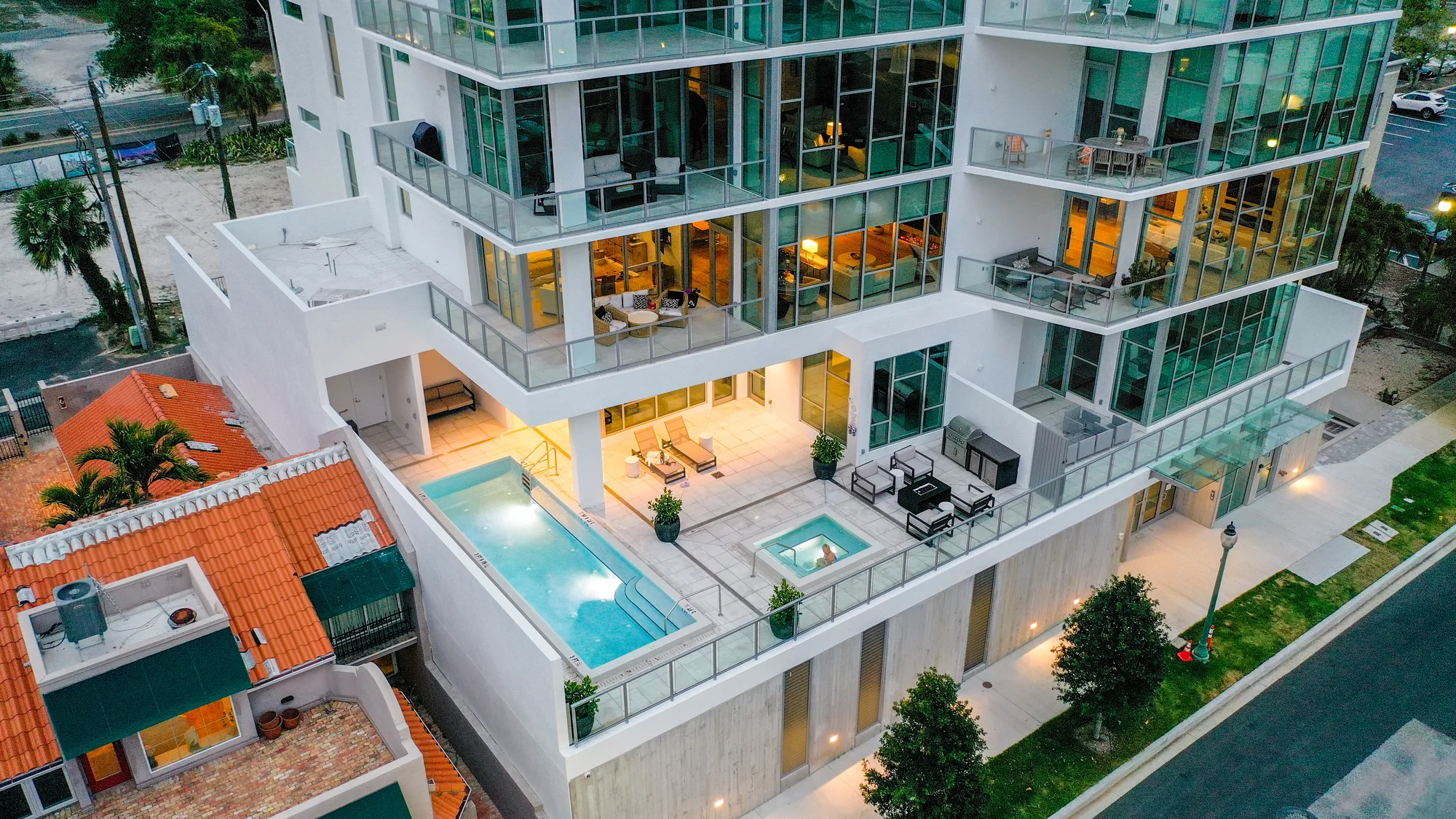 An aerial view of a contemporary apartment building featuring large glass windows and two swimming pools on the ground level. The surrounding area includes landscaped greenery and nearby residential rooftops.
