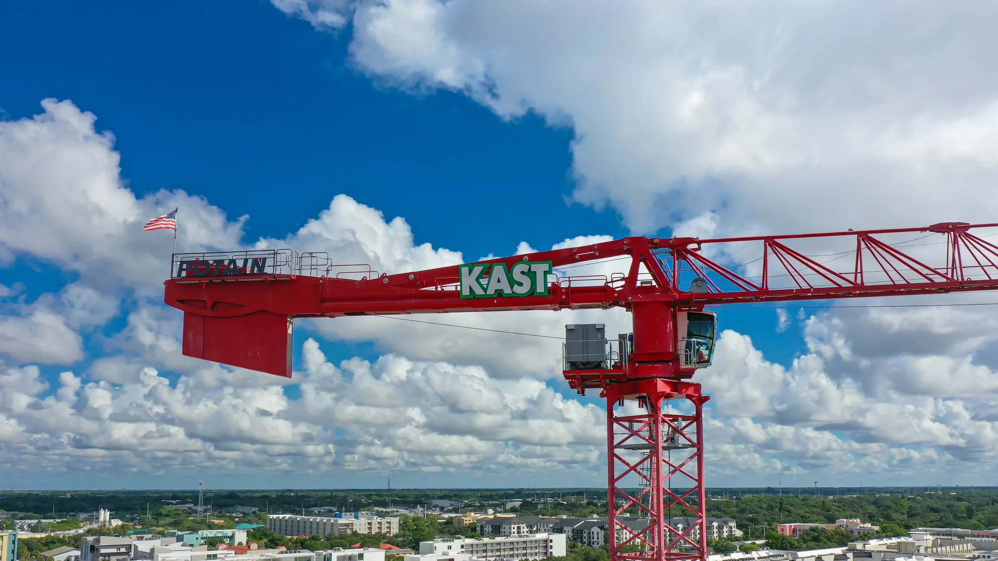 A large red construction crane stands tall against a blue sky filled with clouds, featuring an American flag waving on top. The crane is labeled with 'KAST' and overlooks a cityscape below.