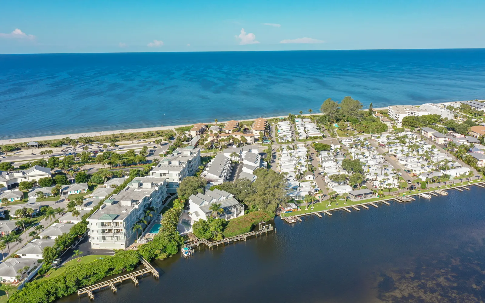 An aerial view showcasing a coastal community adjacent to a beach and the ocean. The scene features residential buildings, a marina, and lush greenery along the shoreline.