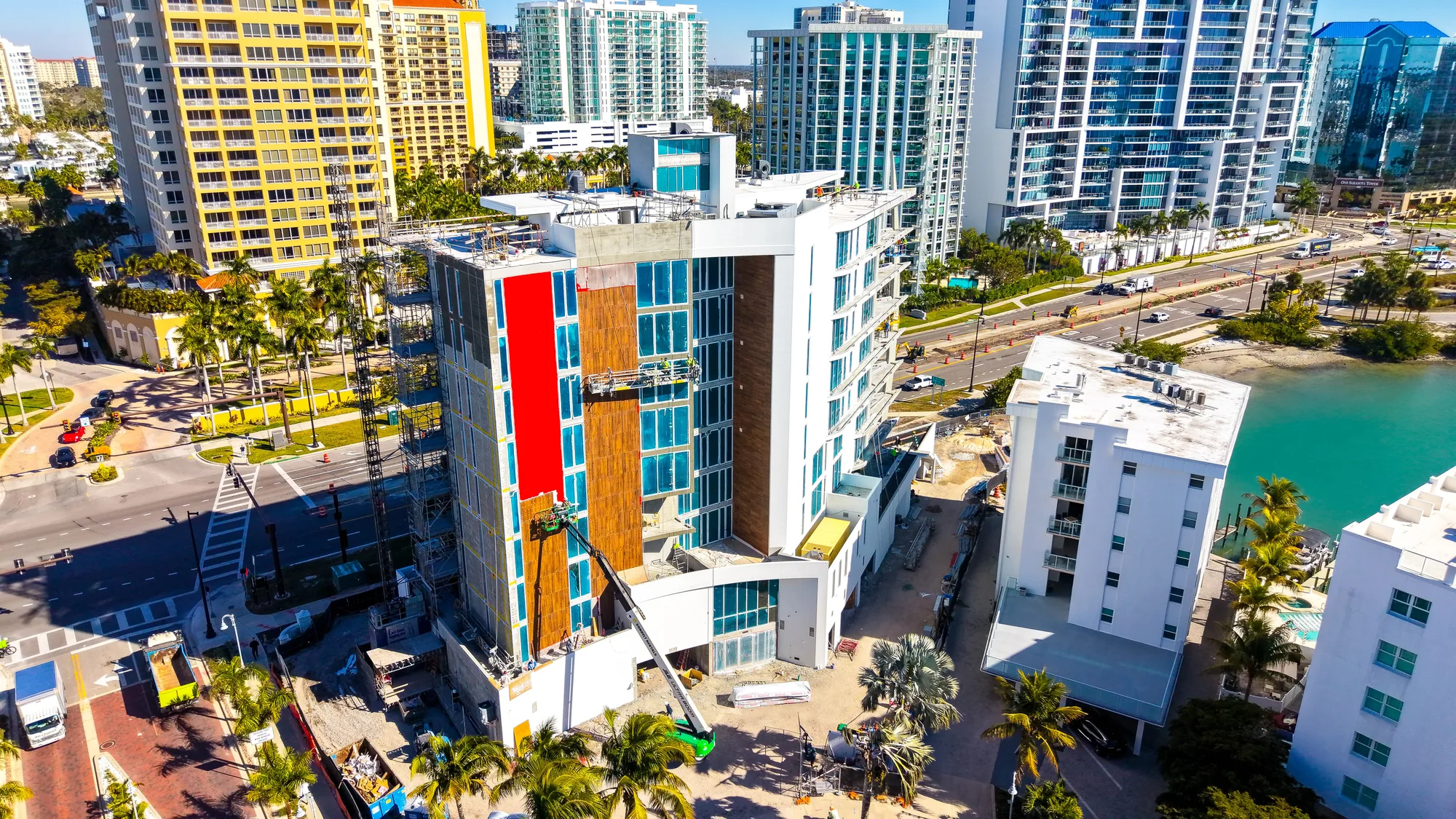 The image shows a modern building under construction, featuring scaffolding and workers on the exterior. Surrounding the site are residential and commercial high-rise buildings, with a view of a roadway and palm trees in the foreground.
