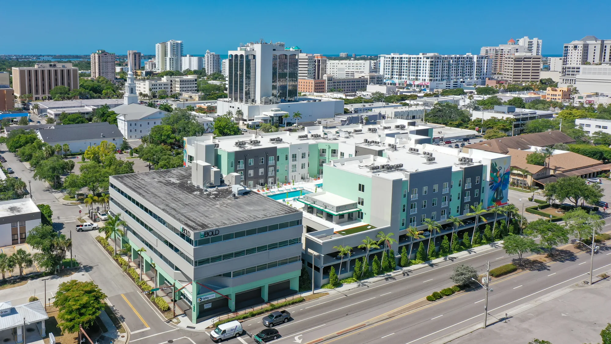 The image showcases an aerial view of a modern urban complex featuring an apartment building with a swimming pool, surrounded by various other buildings and greenery. In the background, the city skyline and clear blue skies are visible, indicating a vibrant metropolitan area.