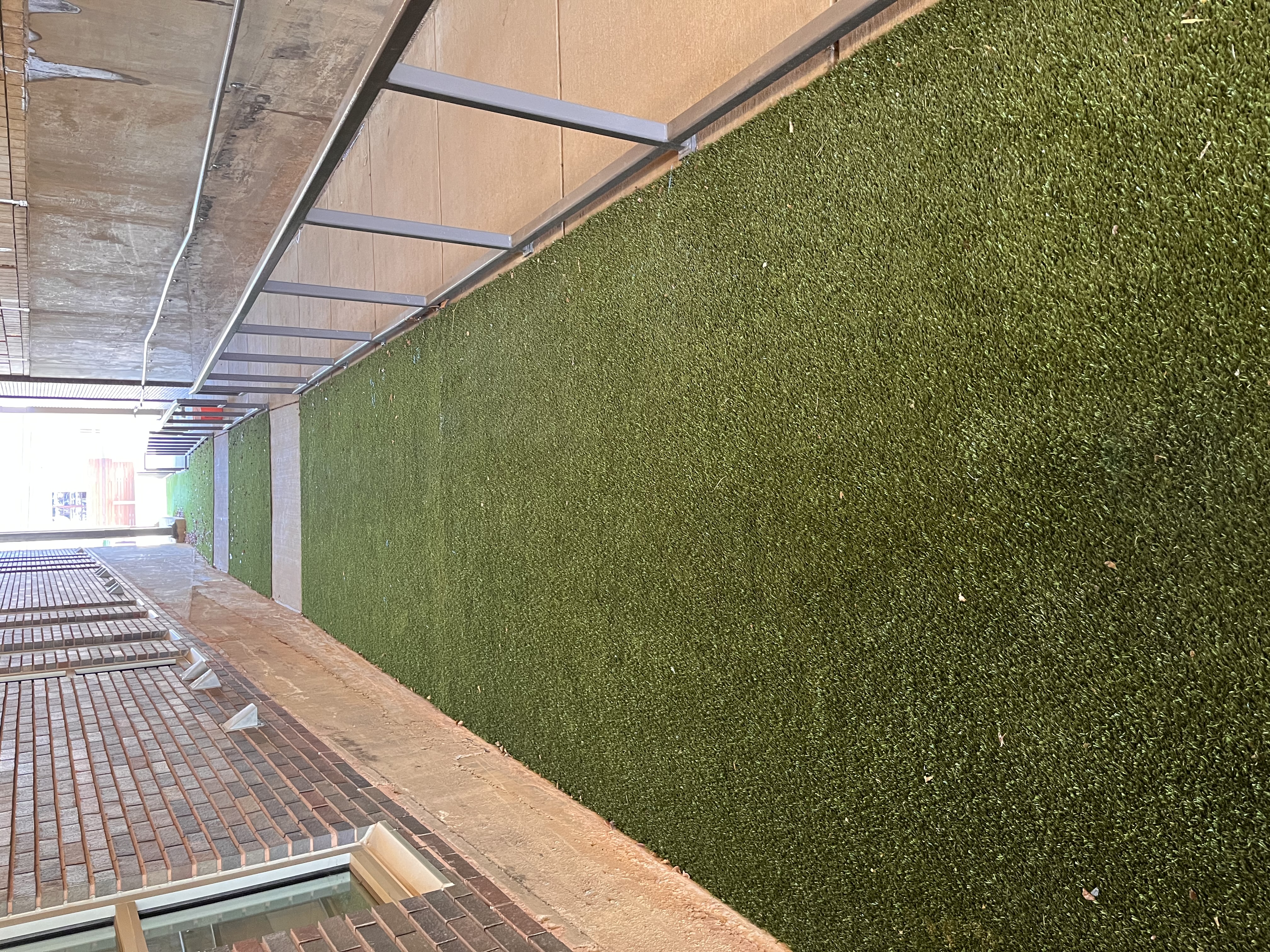 Narrow artificial turf pathway between brick walls with a railing on one side leading towards an open area