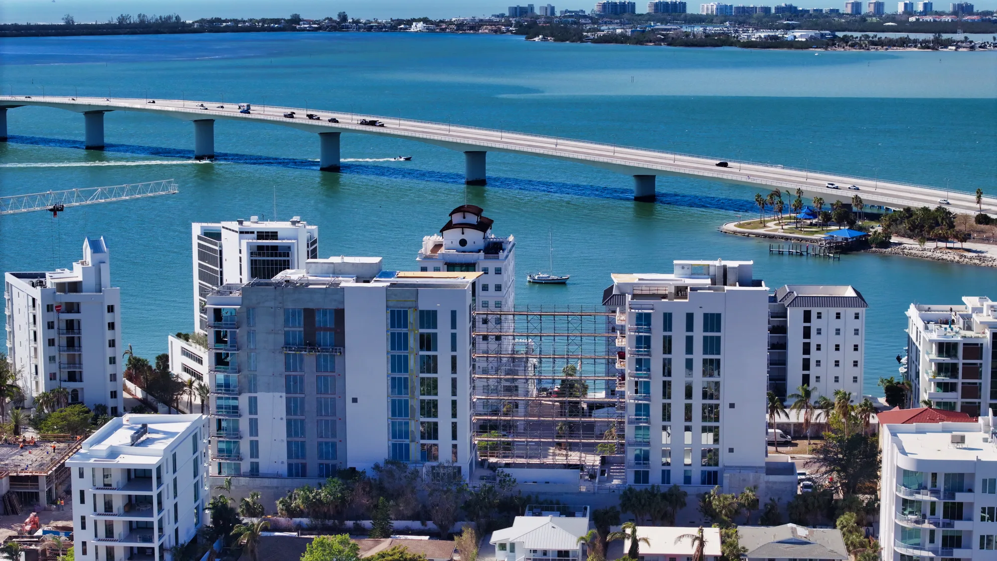 An aerial view of a coastal city featuring modern buildings and a bridge spanning across turquoise waters. A small boat is seen near the shore, with a backdrop of palm trees and other urban structures.
