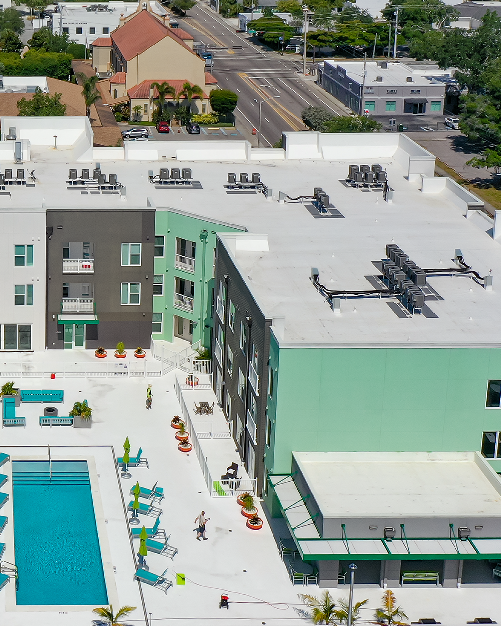 The image shows an aerial view of a modern apartment complex with a swimming pool, surrounded by colorful lounge chairs and potted plants. Nearby, residential and commercial buildings, along with a road, are visible in the background.