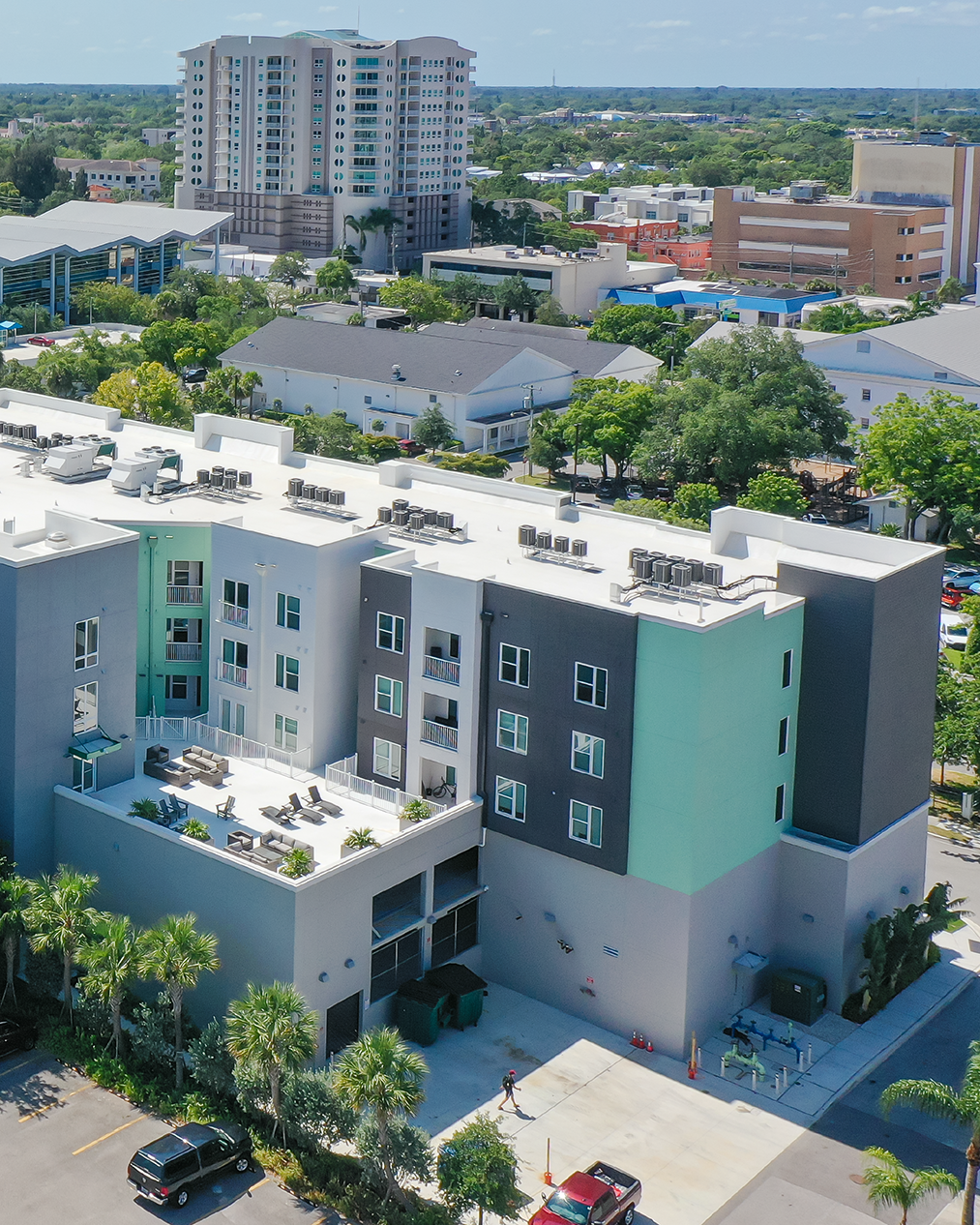 The image shows a modern apartment complex featuring a mix of colors and rooftop seating areas. In the background, a high-rise building and nearby commercial structures can be seen, set among lush greenery.