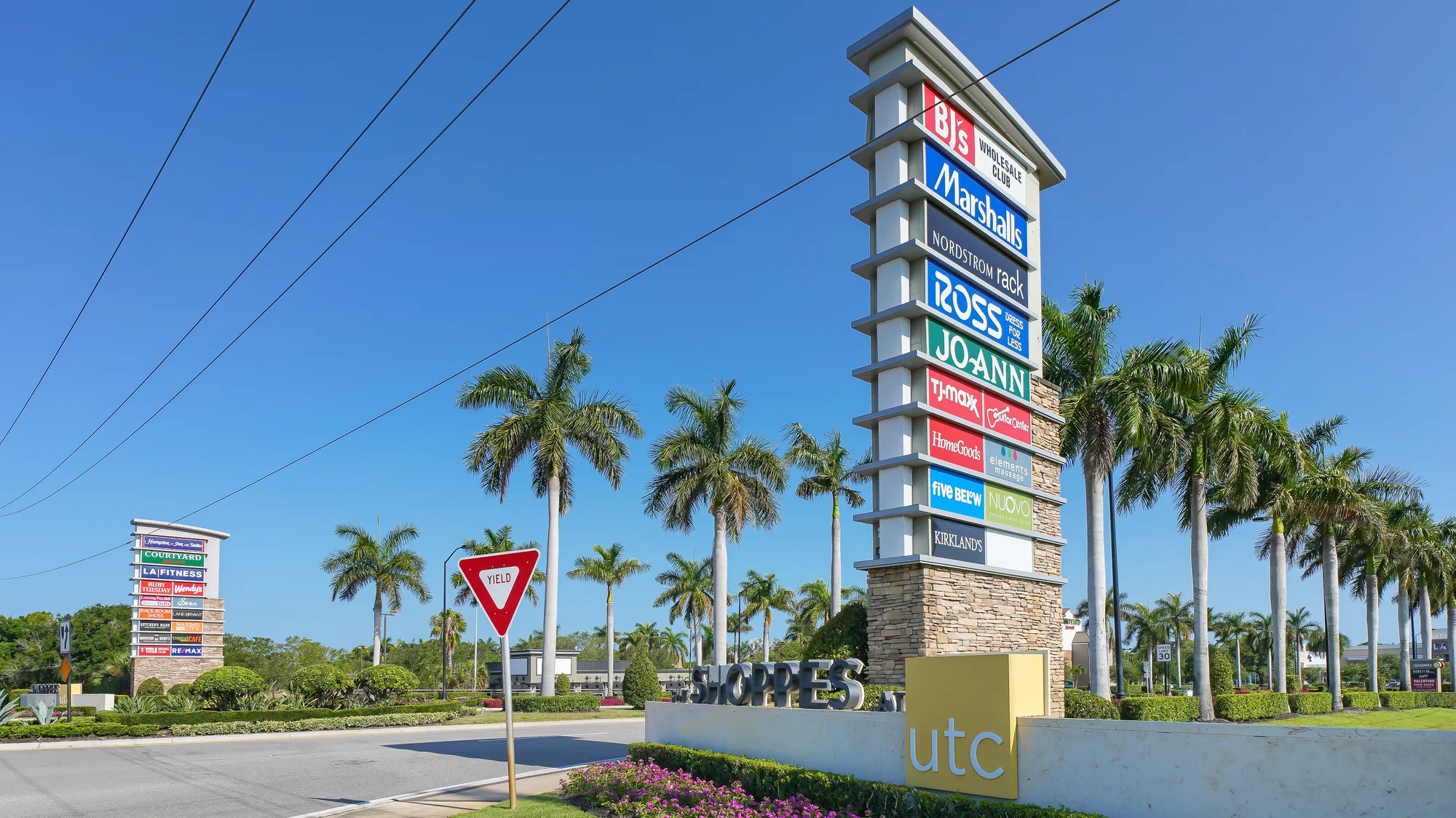 A large, multilevel sign displays various retail store names, including BJ's, Marshalls, and TJ Maxx, at a shopping center. The scene is accompanied by palm trees and a clear blue sky, indicating a sunny day.