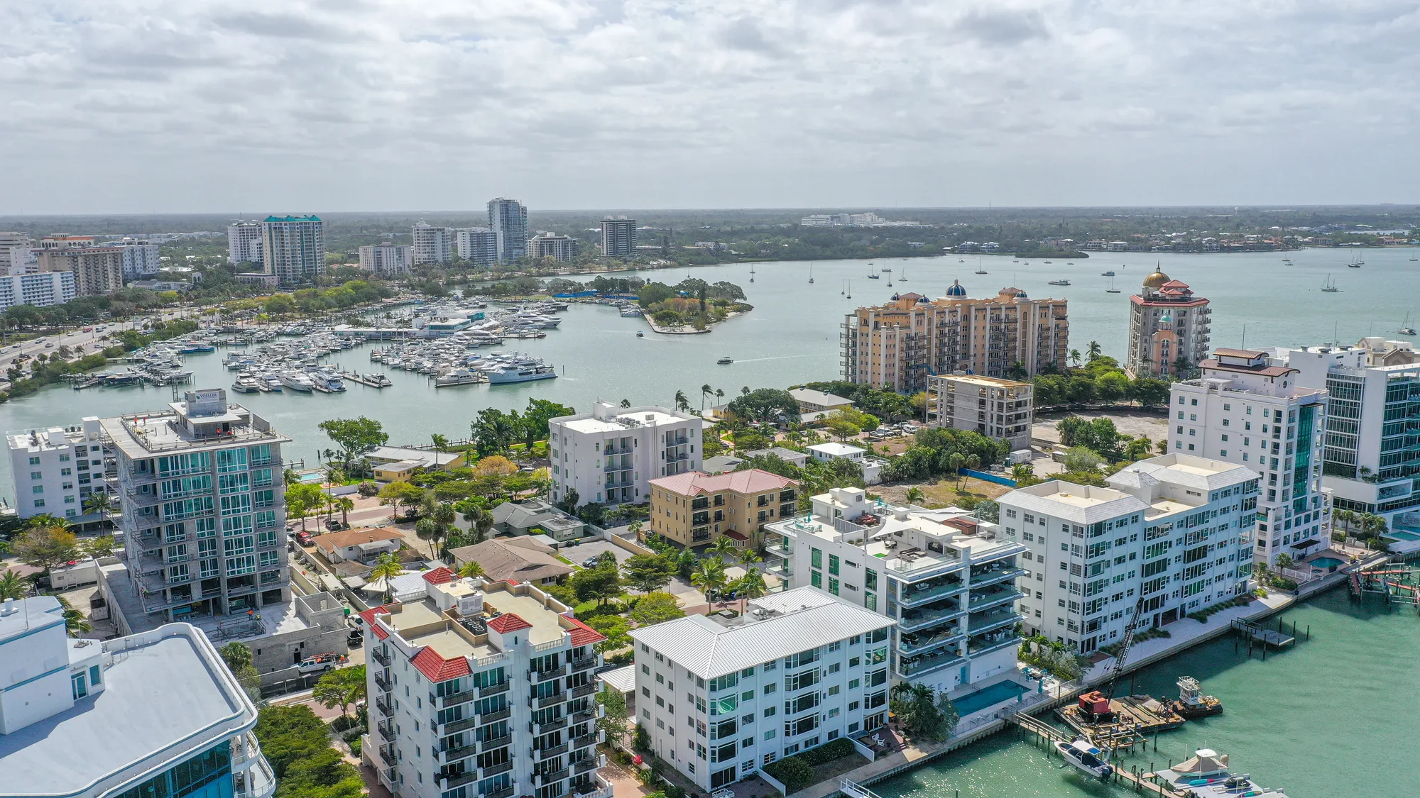 An aerial view showcasing a vibrant coastal city with residential buildings lining the waterfront. A marina filled with boats is visible in the background, and lush greenery complements the urban landscape.