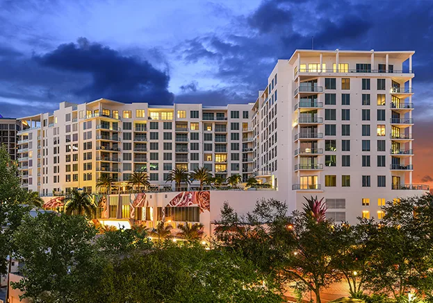 A contemporary multi-story apartment building featuring large windows and balconies, surrounded by lush landscaping. The sky is illuminated by twilight, highlighting the building's architectural design.