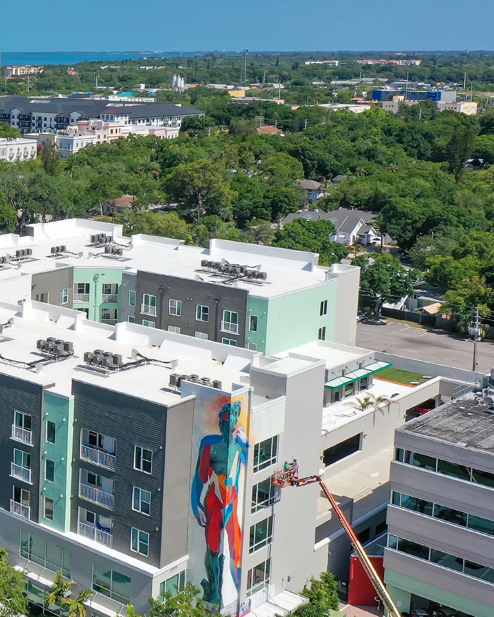 An aerial view of a modern building featuring a vibrant mural on its exterior, depicting a colorful figure. A cherry picker is seen with artists adding finishing touches to the artwork, surrounded by lush greenery and a cityscape in the background.