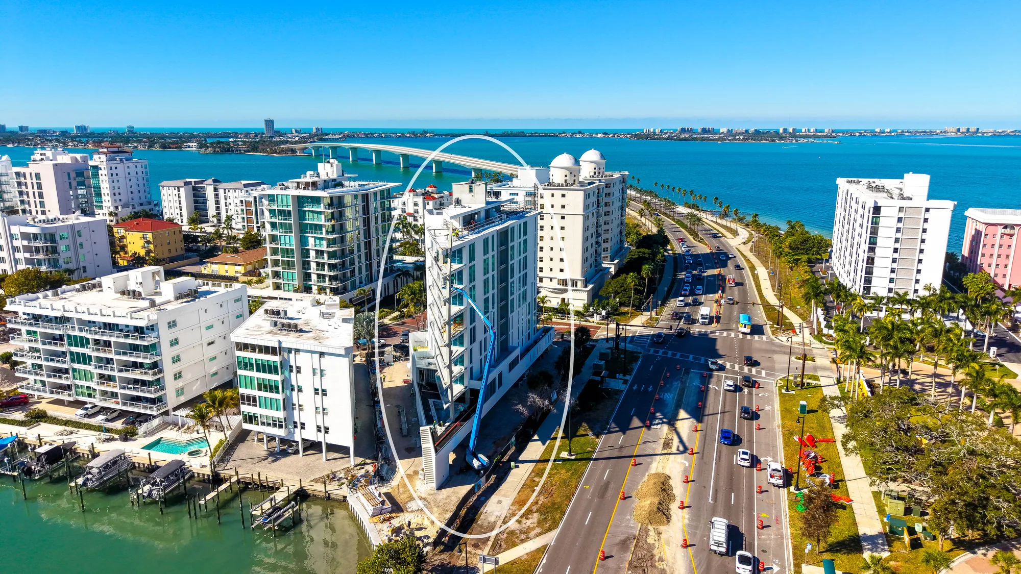 An aerial view of a coastal city featuring modern high-rise buildings along a waterway, with construction work visible on one of the structures. The scene includes vehicles on a roadway, palm trees, and a clear blue sky, highlighting a vibrant urban environment.