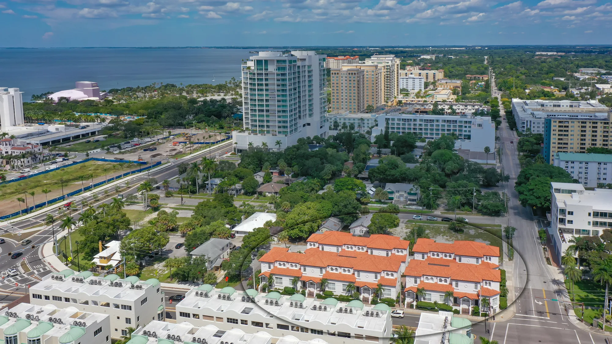 The image captures an aerial view of a coastal city featuring a mix of residential and commercial buildings, along with lush greenery. In the background, the expansive waterfront can be seen, flanked by various high-rise structures and developments.