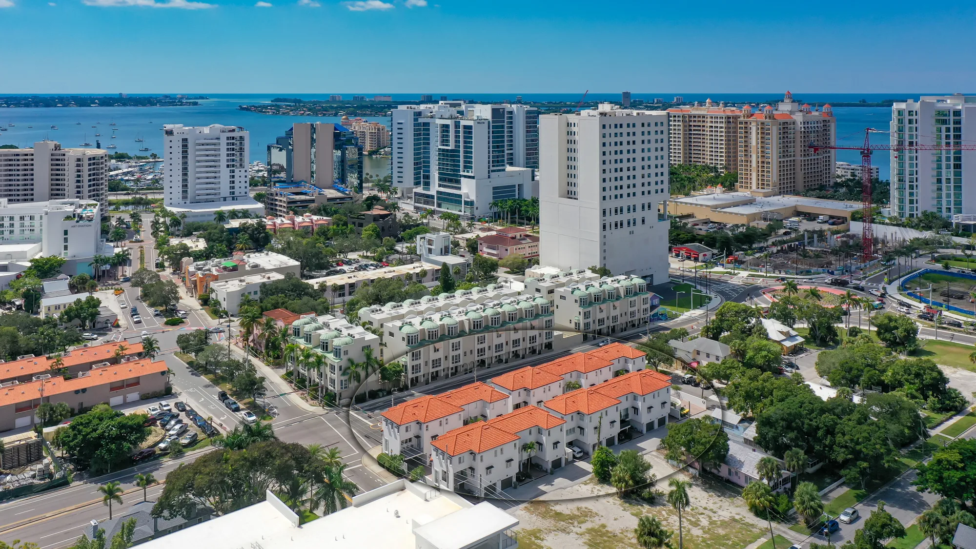 This aerial image showcases a vibrant urban landscape with a mix of high-rise buildings and residential areas along a waterfront. Several boats are visible in the water, and lush greenery intersperses with busy streets, highlighting a mix of development and nature.