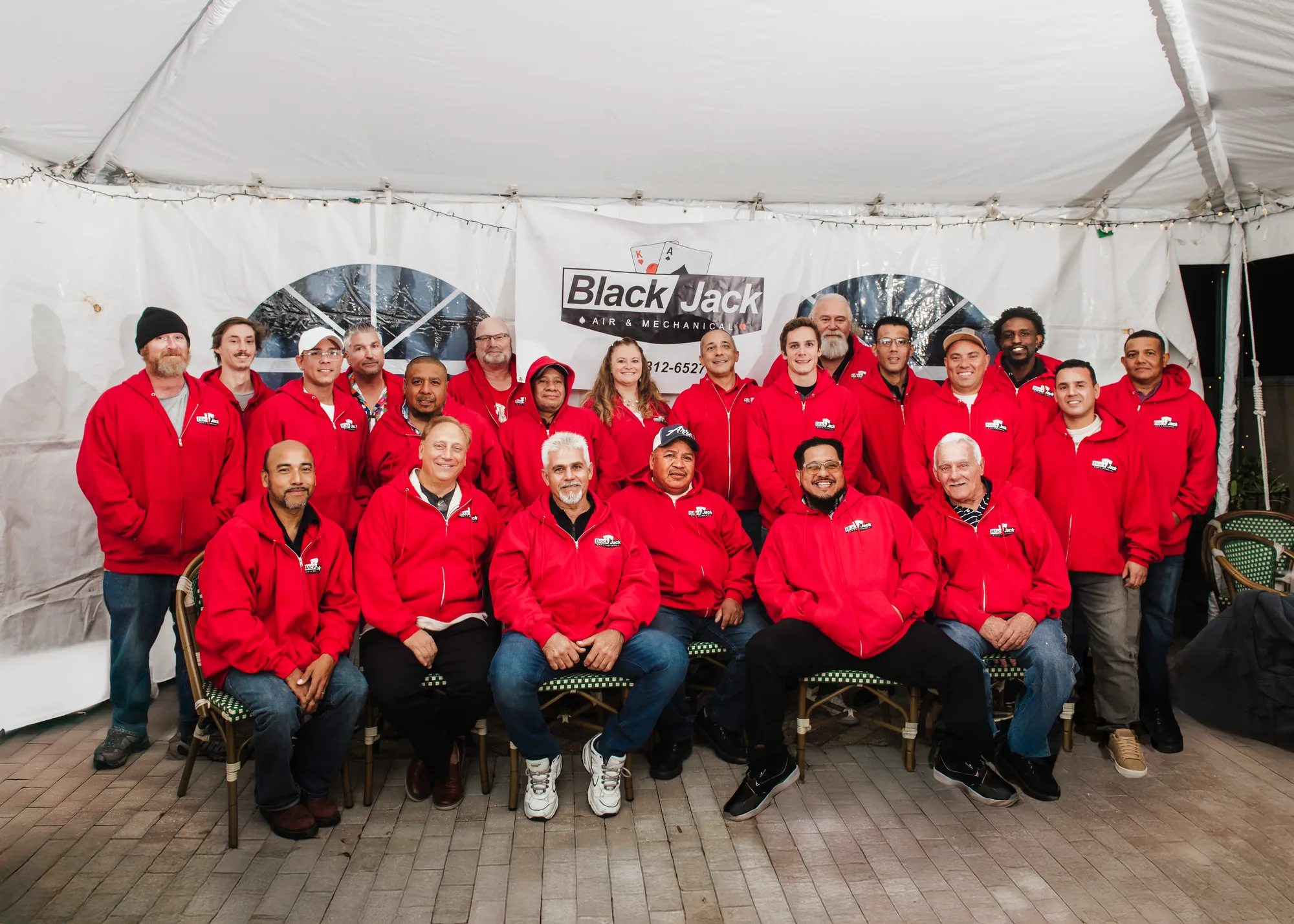 A group photo featuring the team from Black Jack Air & Mechanical, dressed in matching red hoodies. They are gathered under a tent with a company banner in the background, showcasing camaraderie and teamwork.