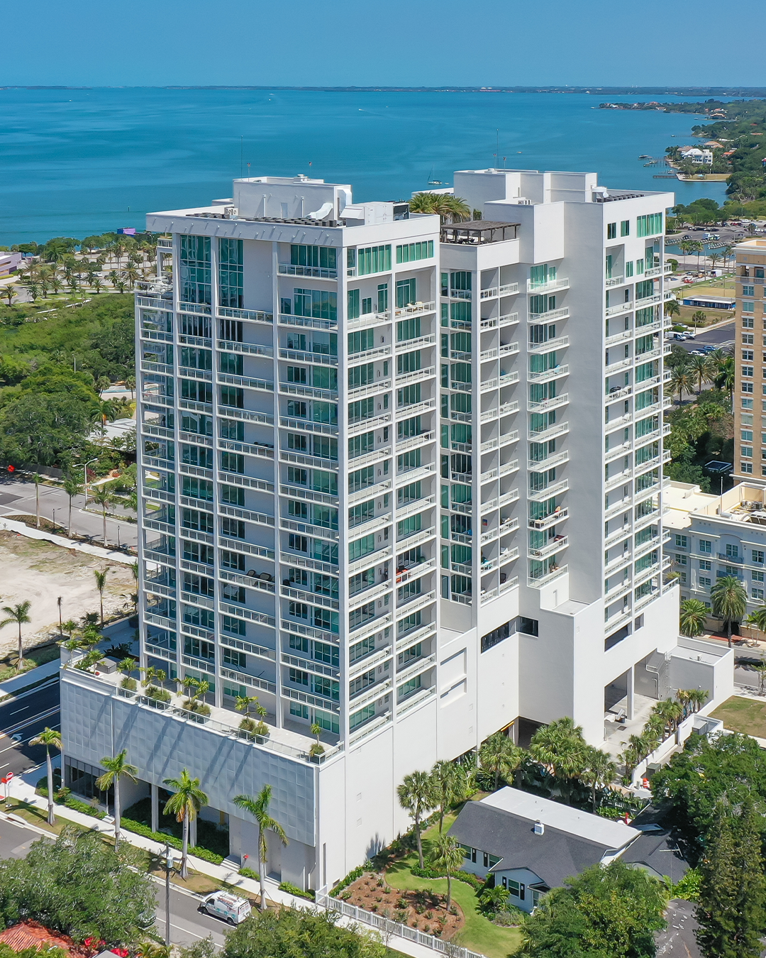 The image showcases a modern, multi-story residential building with large glass windows, situated near a coastline. In the background, the turquoise water and green landscape highlight the building's scenic location.