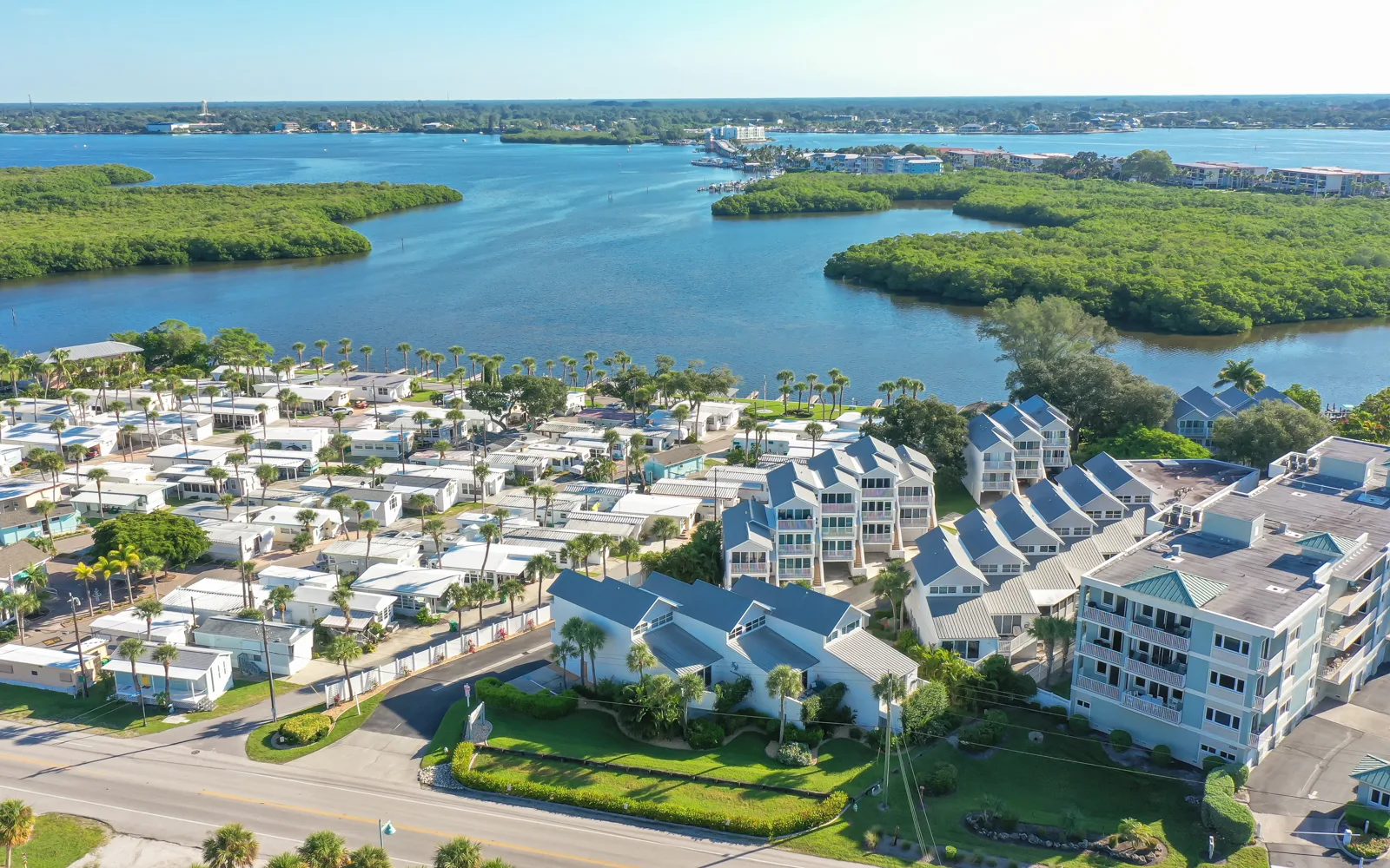 This image captures an aerial view of a coastal community with residential buildings and mobile homes, situated near a body of water surrounded by lush greenery. The tranquil setting features palm trees and a clear blue sky, highlighting the natural beauty of the area.