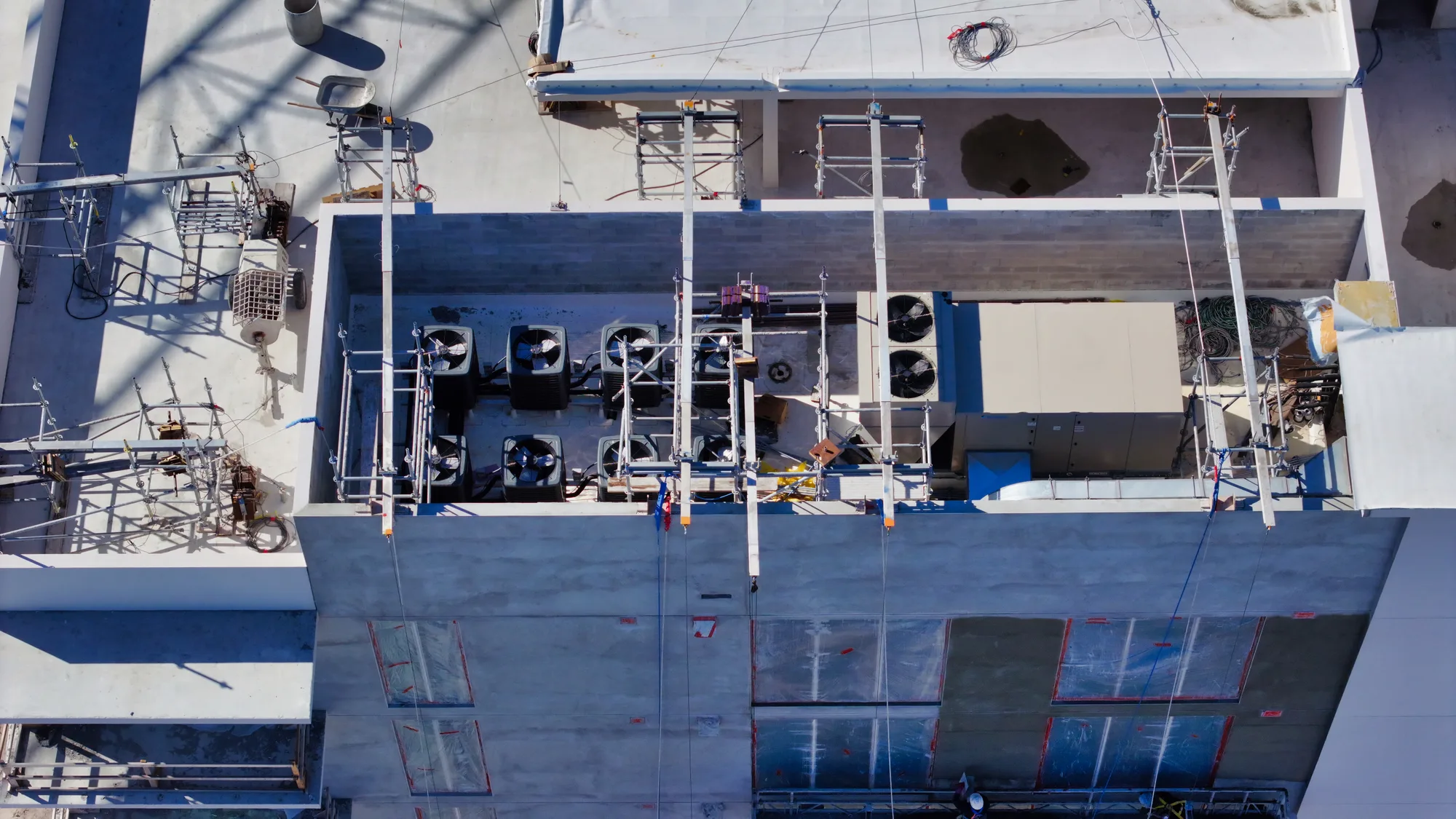 An aerial view of a construction site rooftop displaying scaffolding, cooling units, and various construction materials. The site features several air conditioning units and equipment positioned on the flat roof amidst scaffolding for ongoing construction work.