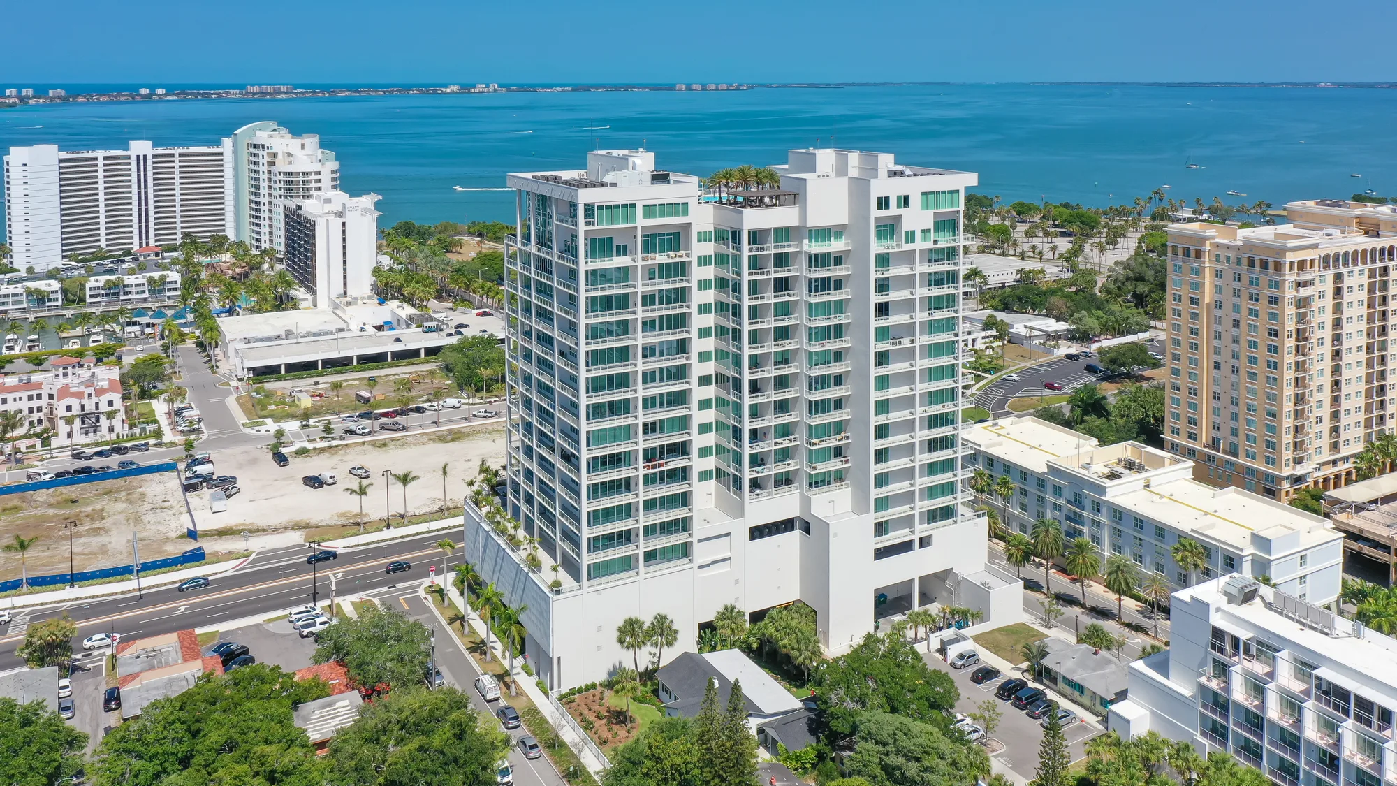 An aerial view of a modern high-rise building situated near the coast, surrounded by palm trees and other structures. The turquoise waters can be seen in the background, highlighting the coastal setting.