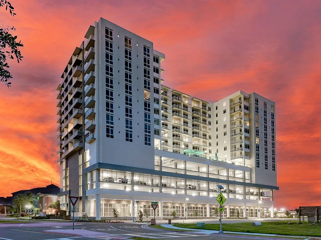 A contemporary multi-story apartment building is showcased against a vibrant sunset, featuring large windows and balconies. The structure appears prominently on a circular intersection, highlighting its modern architectural design.