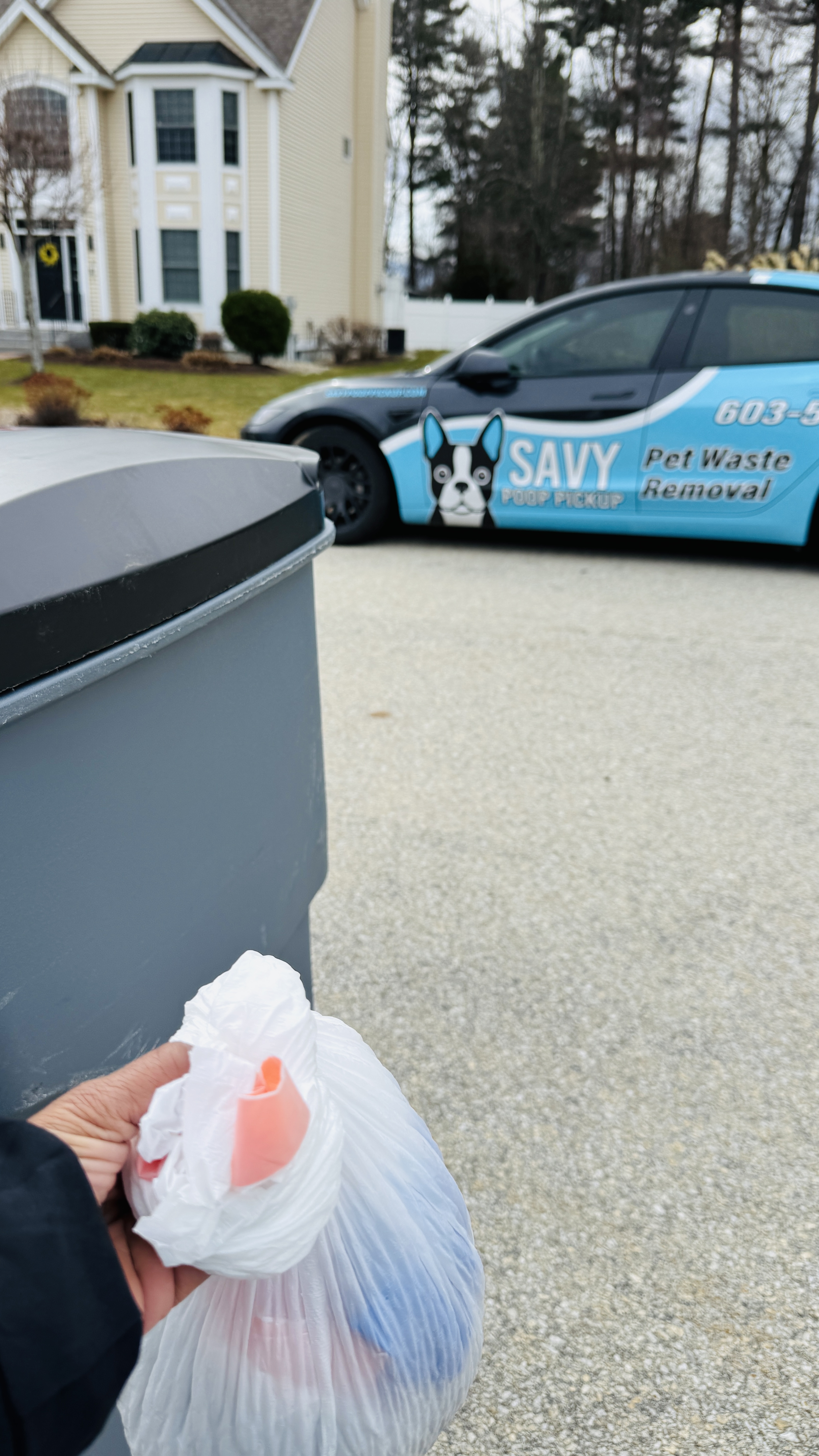 Bagged pet waste being placed in a customer's gray outdoor trash barrel with the Savy vehicle behind