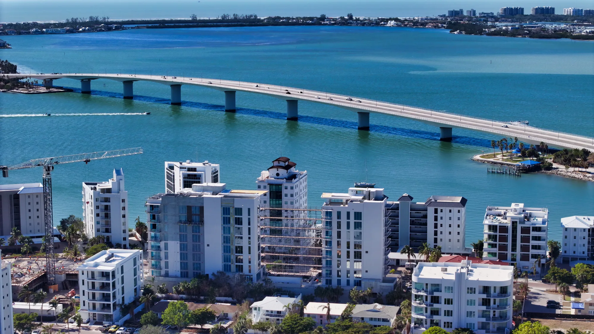An aerial view showcasing a coastal city with modern buildings, a construction site, and a curved bridge crossing over blue waters. Several boats can be seen navigating the water, contributing to the vibrant urban landscape.