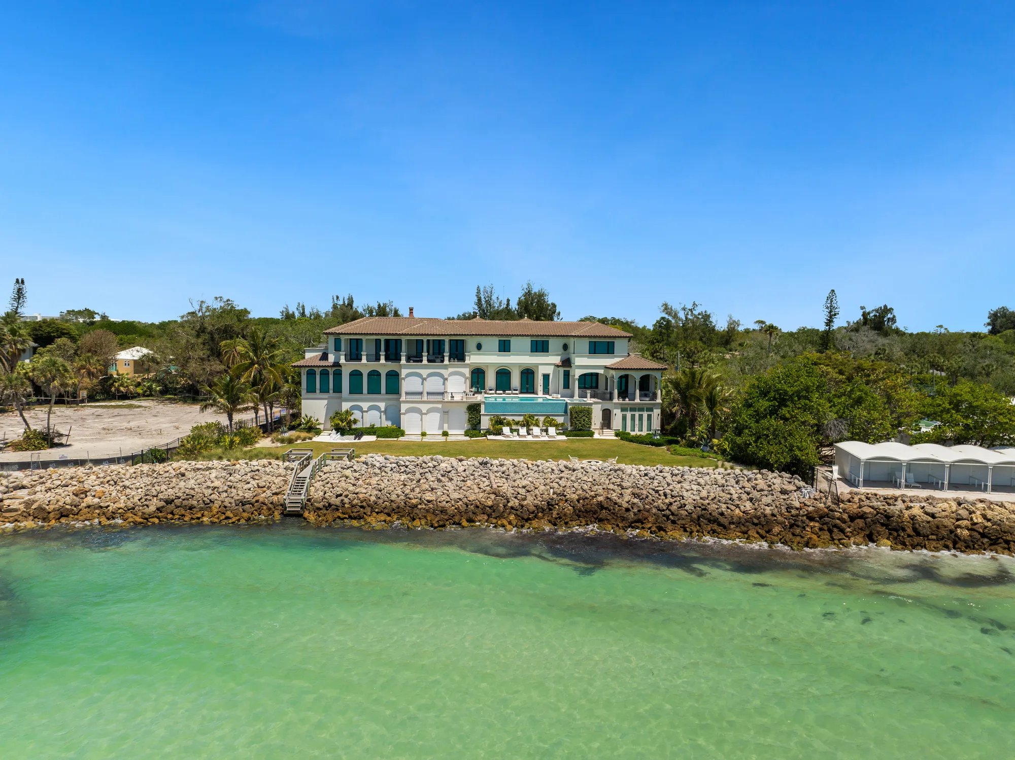 A stunning waterfront mansion features a blend of greenery and rocky shoreline. The spacious property boasts large windows, a pool area, and palm trees, situated against a clear blue sky.